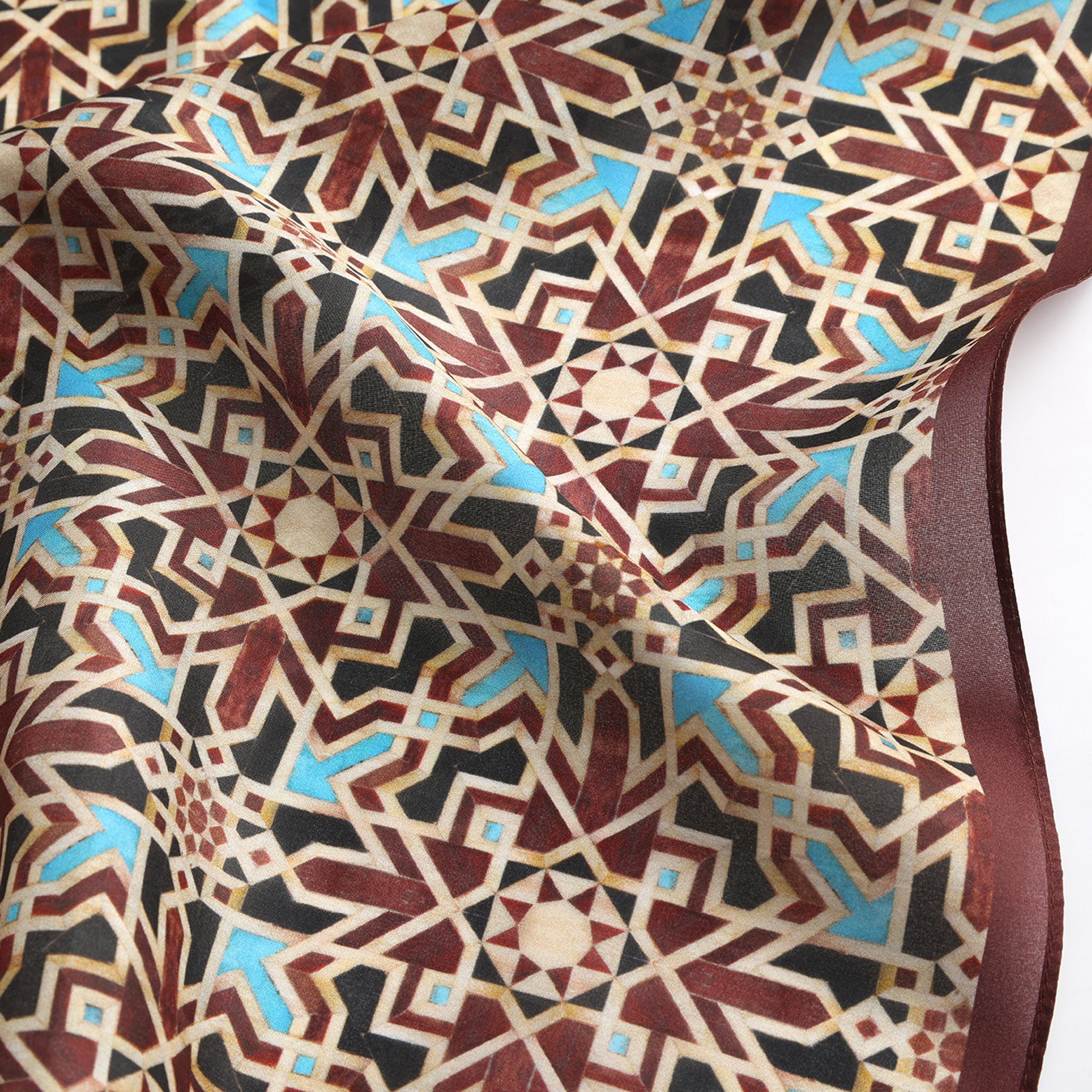 The Brown Silk Neckerchief showcases intricate geometric patterns with interlocking stars and shapes in brown, beige, and blue hues, inspired by Islamic art. Its soft texture reflects Spanish craftsmanship.