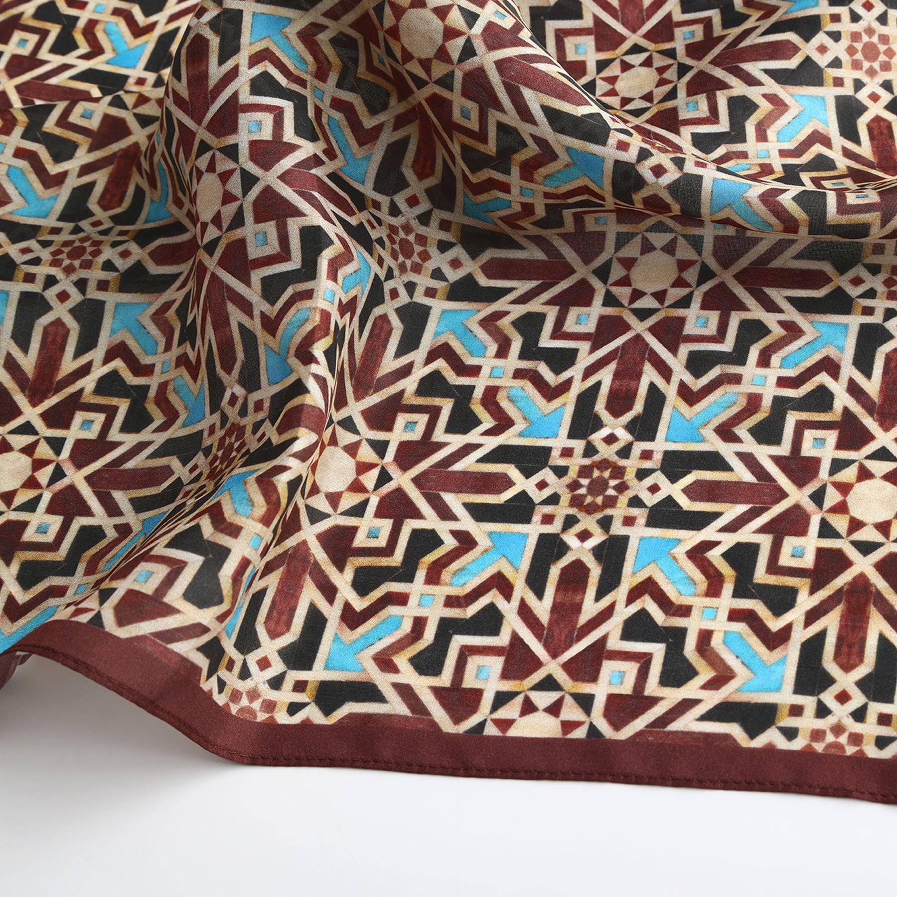 The Brown Silk Neckerchief features an Islamic art-inspired geometric pattern in burgundy, beige, black, and blue, with repeating star motifs and intersecting lines. Its slight drape highlights the fabric's texture and sheen.