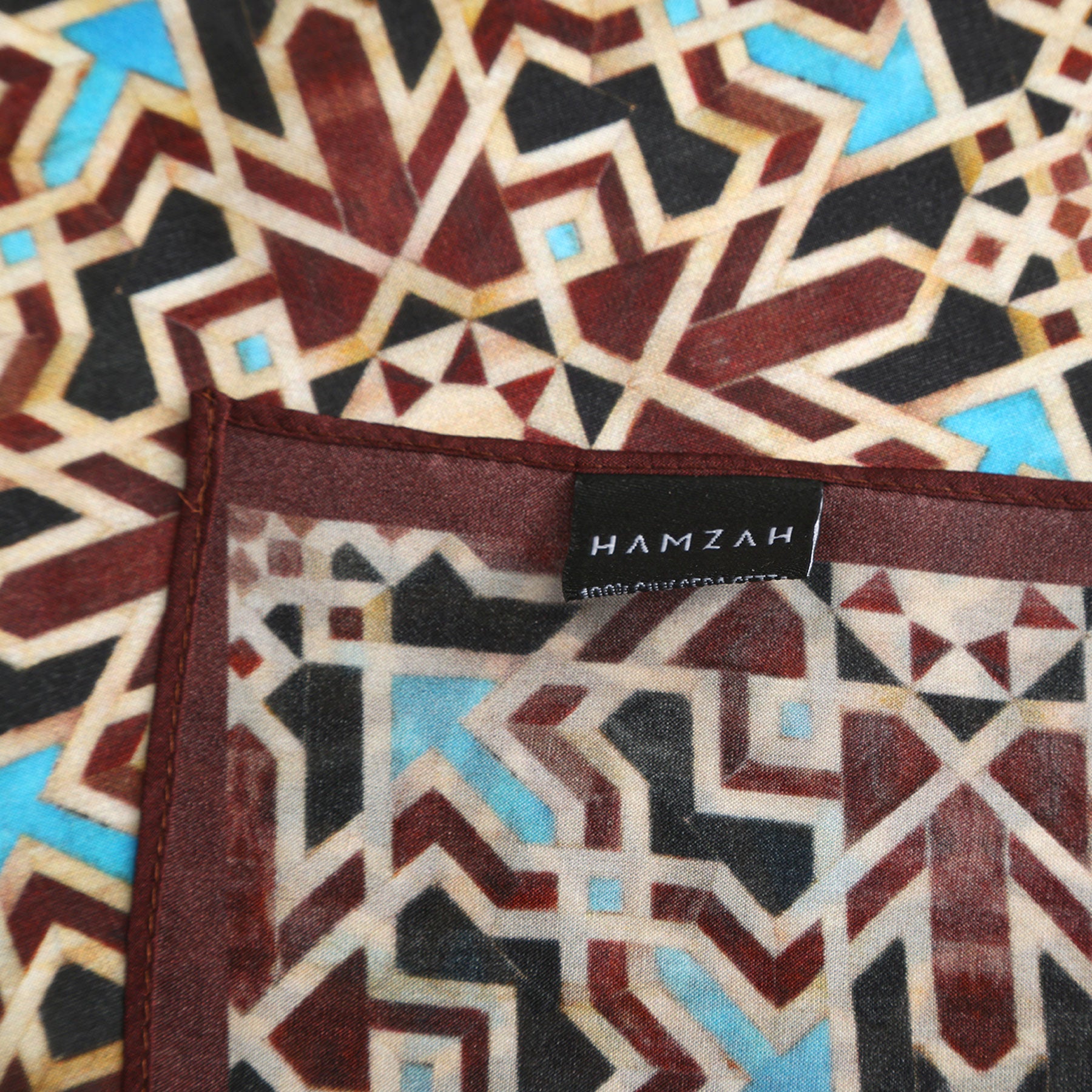 A Brown Silk Neckerchief with an intricate geometric Islamic art print in brown, beige, and blue, featuring a black sewn-on “HAMZAH” label in white text.
