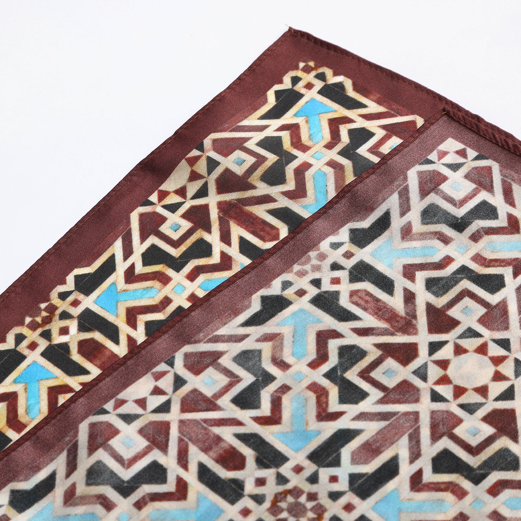 Two corners of the Brown Silk Neckerchief, featuring intricate geometric patterns in brown, blue, cream, and black with a maroon border, displayed against a white background—a tribute to Spanish craftsmanship.