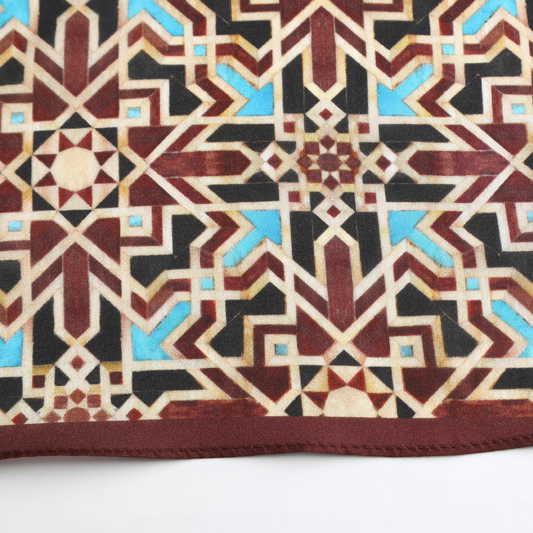 A close-up of the Brown Silk Neckerchief showcasing a geometric Islamic-inspired print in brown, beige, blue, and maroon, with intersecting lines and stars, finished with a maroon border.