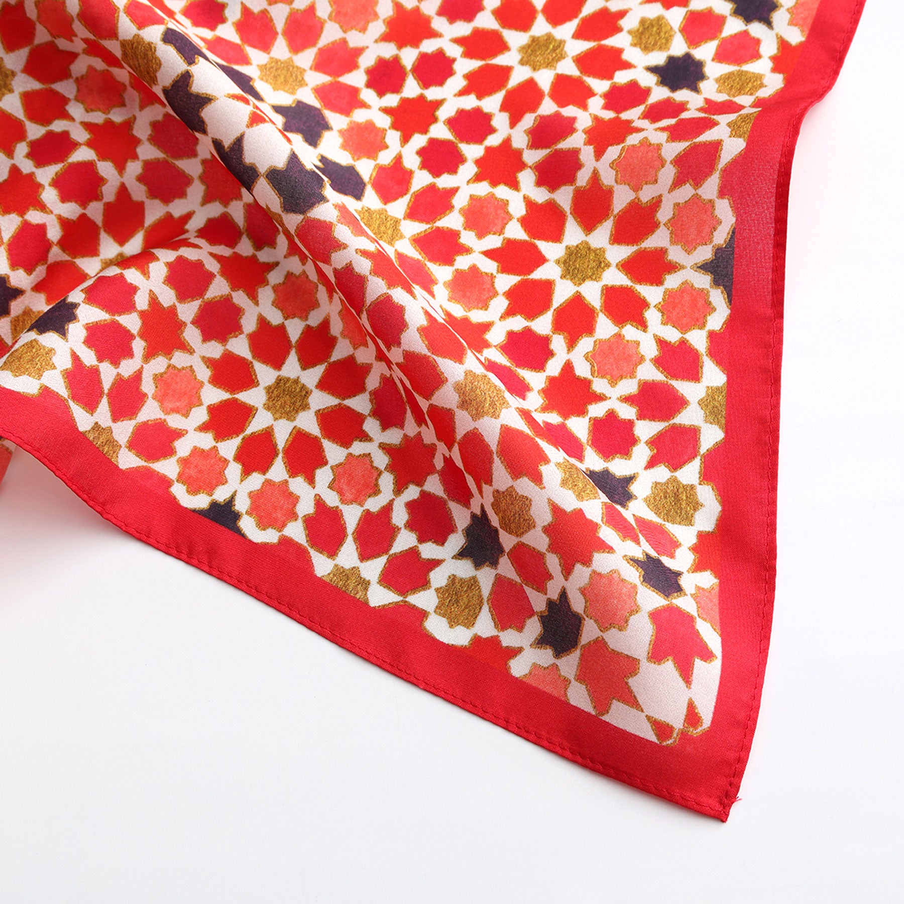 The Small Square Scarf Alhambra is a red-bordered silk square featuring geometric star and floral patterns in red, gold, orange, and purple on a white background, inspired by Islamic art. Shown partially folded on a white surface.