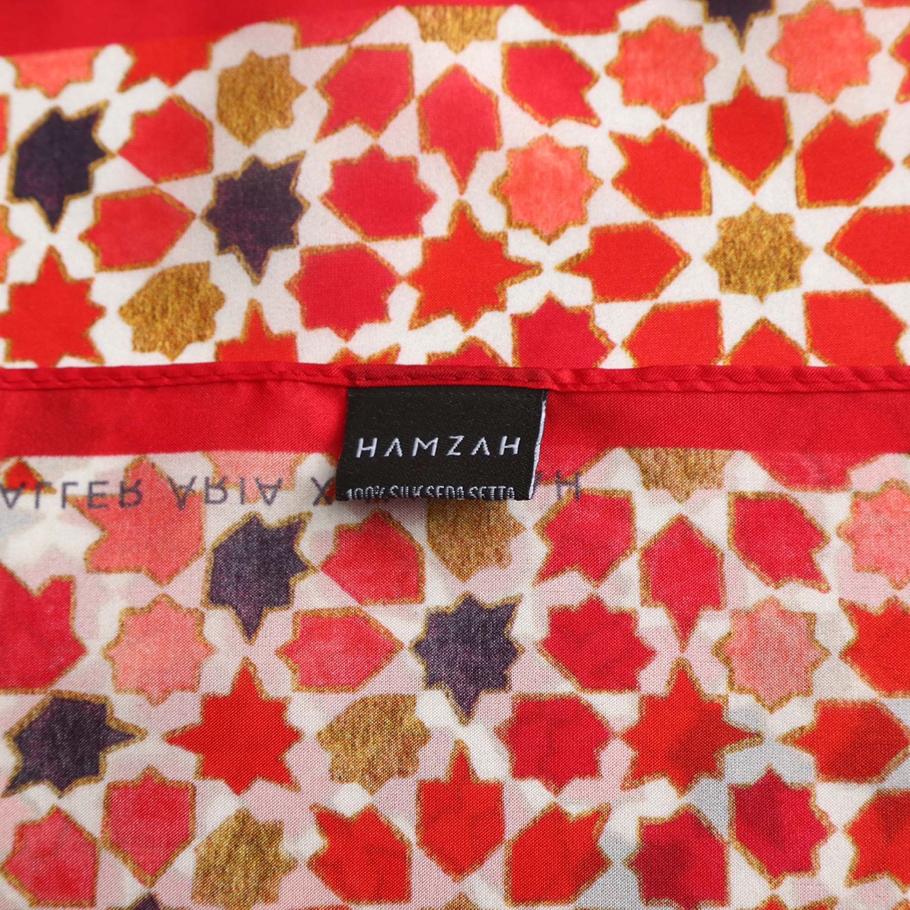 A close-up of the Small Square Scarf Alhambra shows red, gold, and purple geometric star patterns inspired by Islamic art, with a black HAMZAH label stitched onto the 100% SILK SEDA SETA fabric.