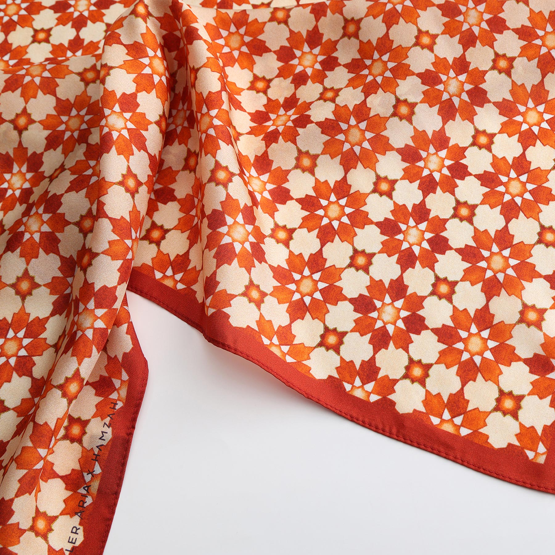 A close-up of the Silk Neckerchief Orange Islamic Art Print shows its intricate geometric star and flower pattern in red, orange, and cream with a solid red edge, part of which is folded over a white surface.