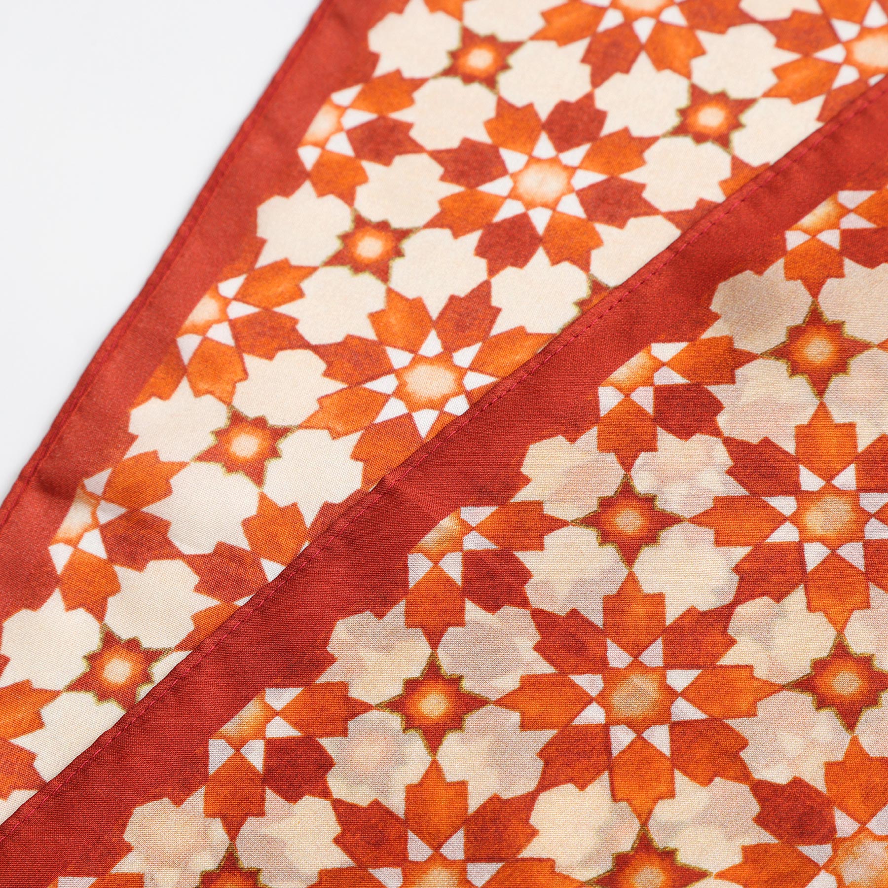 A close-up of the Silk Neckerchief Orange Islamic Art Print highlights Spanish craftsmanship with a geometric, star-inspired pattern in orange, red, and cream. The diagonally folded silk emphasizes the intricate repeating design.