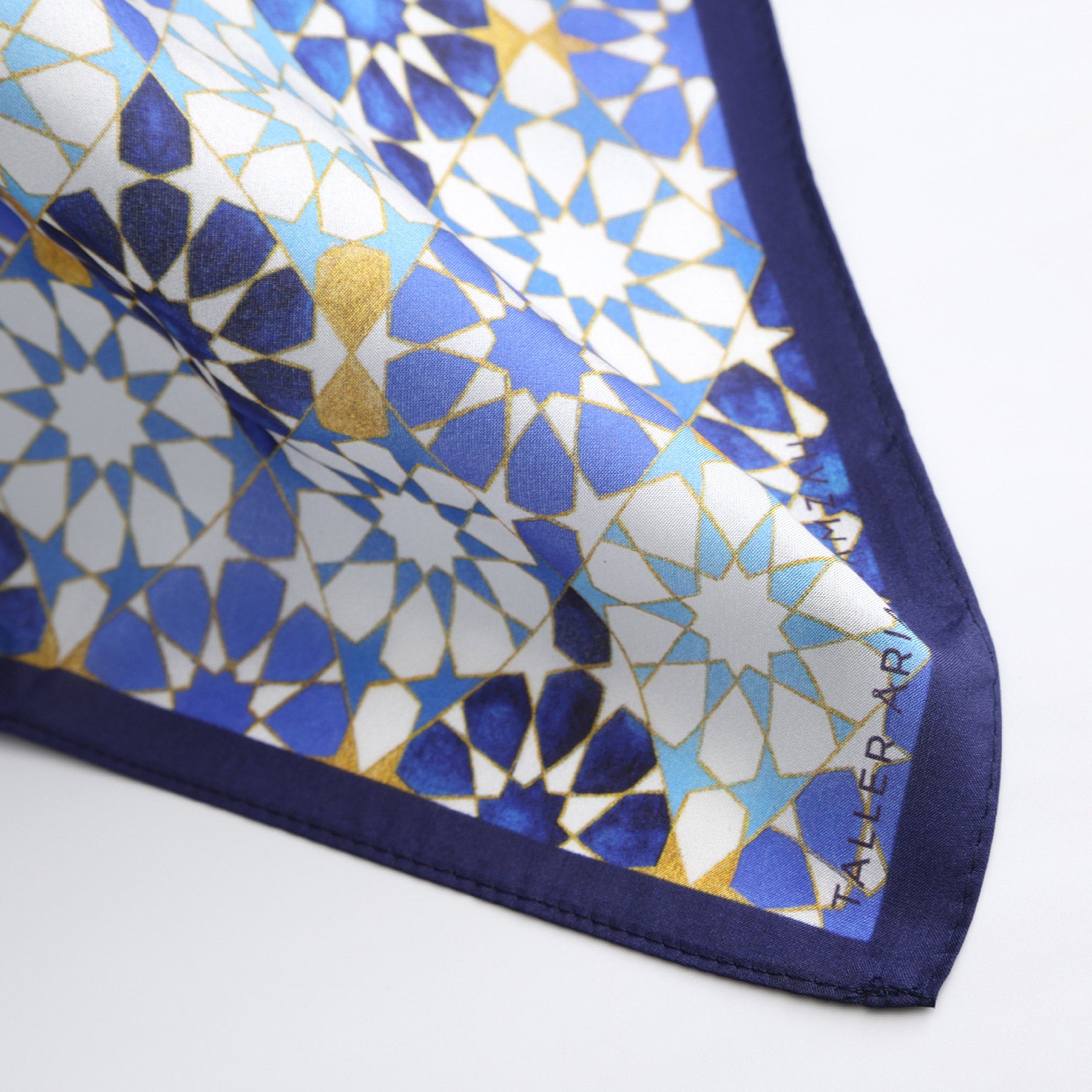 The Blue Neck Scarf 100% Silk features a geometric pattern in blue, white, and gold with a dark blue border, star-like mosaic designs inspired by Islamic art, and partially visible text in the corner.