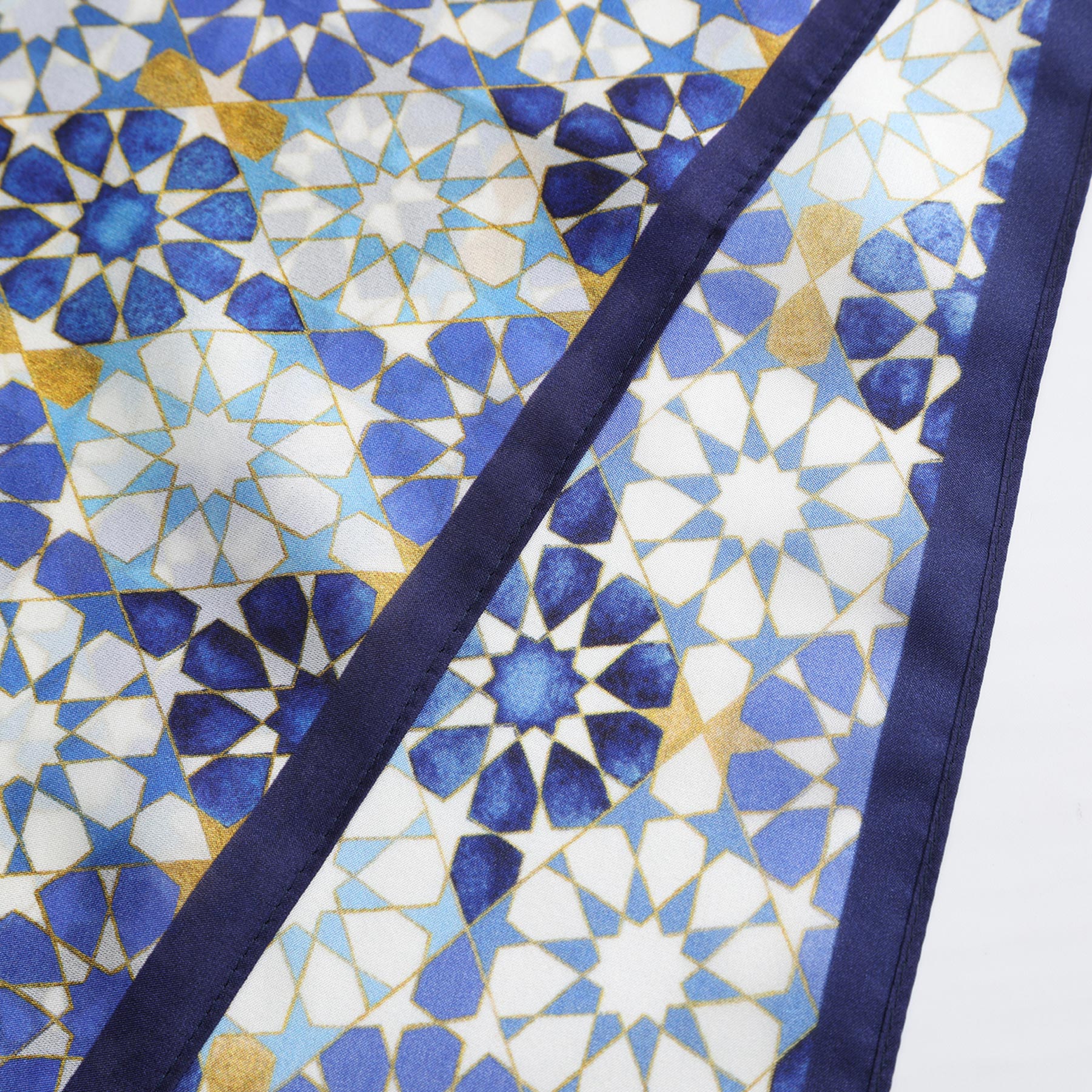 A close-up of the Blue Neck Scarf 100% Silk, featuring an intricate geometric pattern in blue, gold, and white inspired by Islamic art and Alhambra Palace, with star and tile motifs bordered by a solid dark blue edge.
