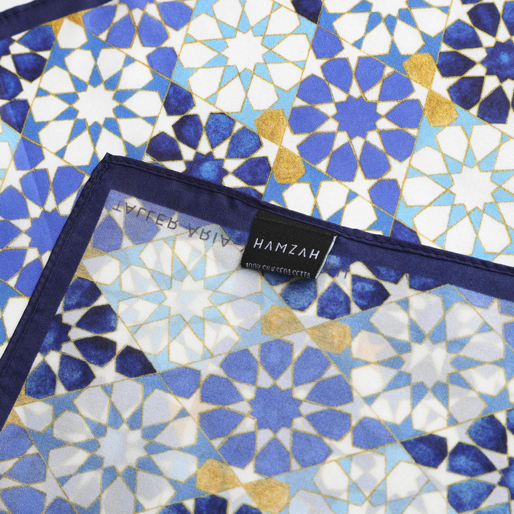 The Blue Neck Scarf 100% Silk features geometric patterns inspired by Islamic art in blue, gold, and white, with a visible “HAMZAH” label and a transparent section showcasing the design on both sides.