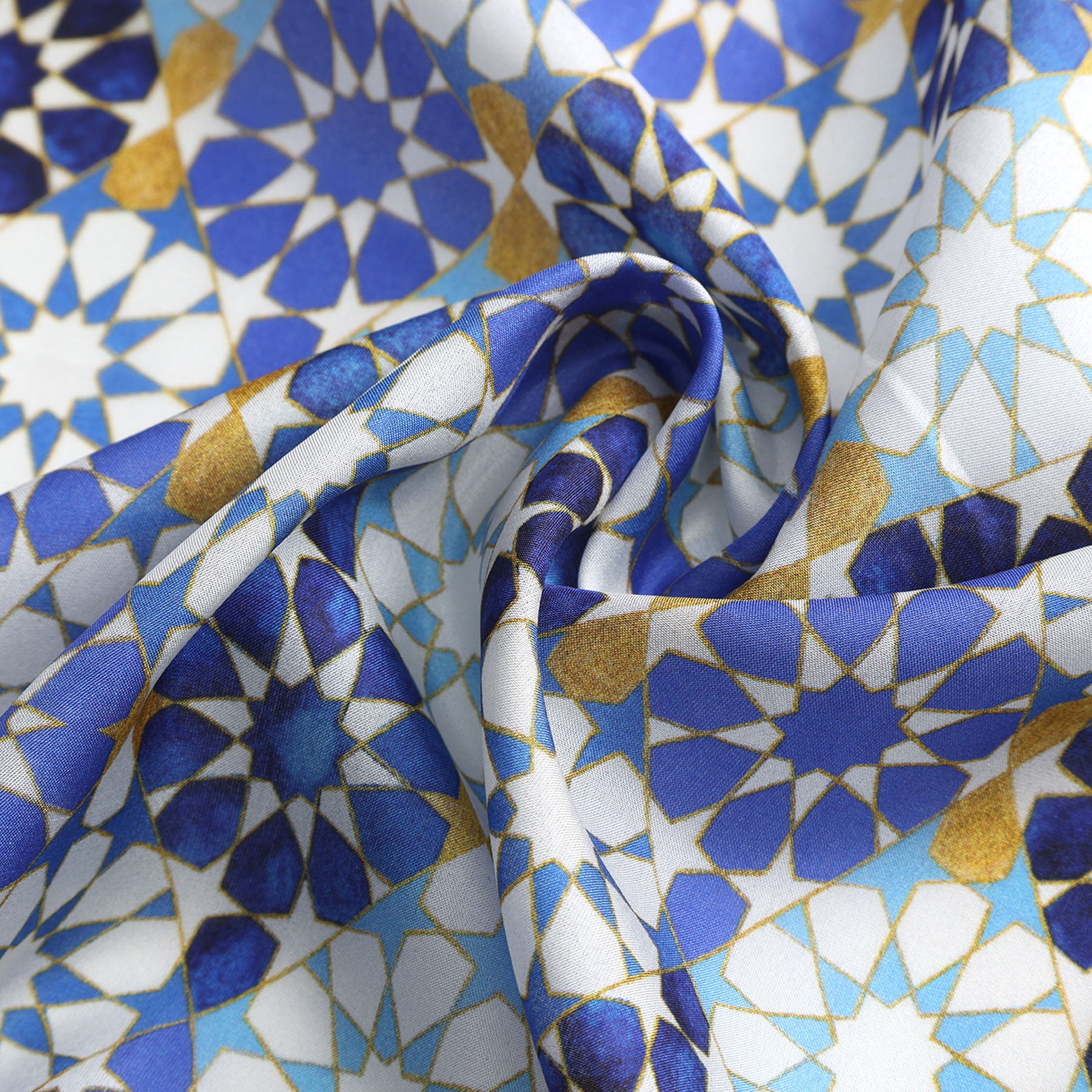 A close-up of the Blue Neck Scarf 100% Silk shows softly folded fabric with a geometric pattern in blue, gold, and white, inspired by Alhambra Palace and Islamic art motifs. The folds create gentle curves and shadows.