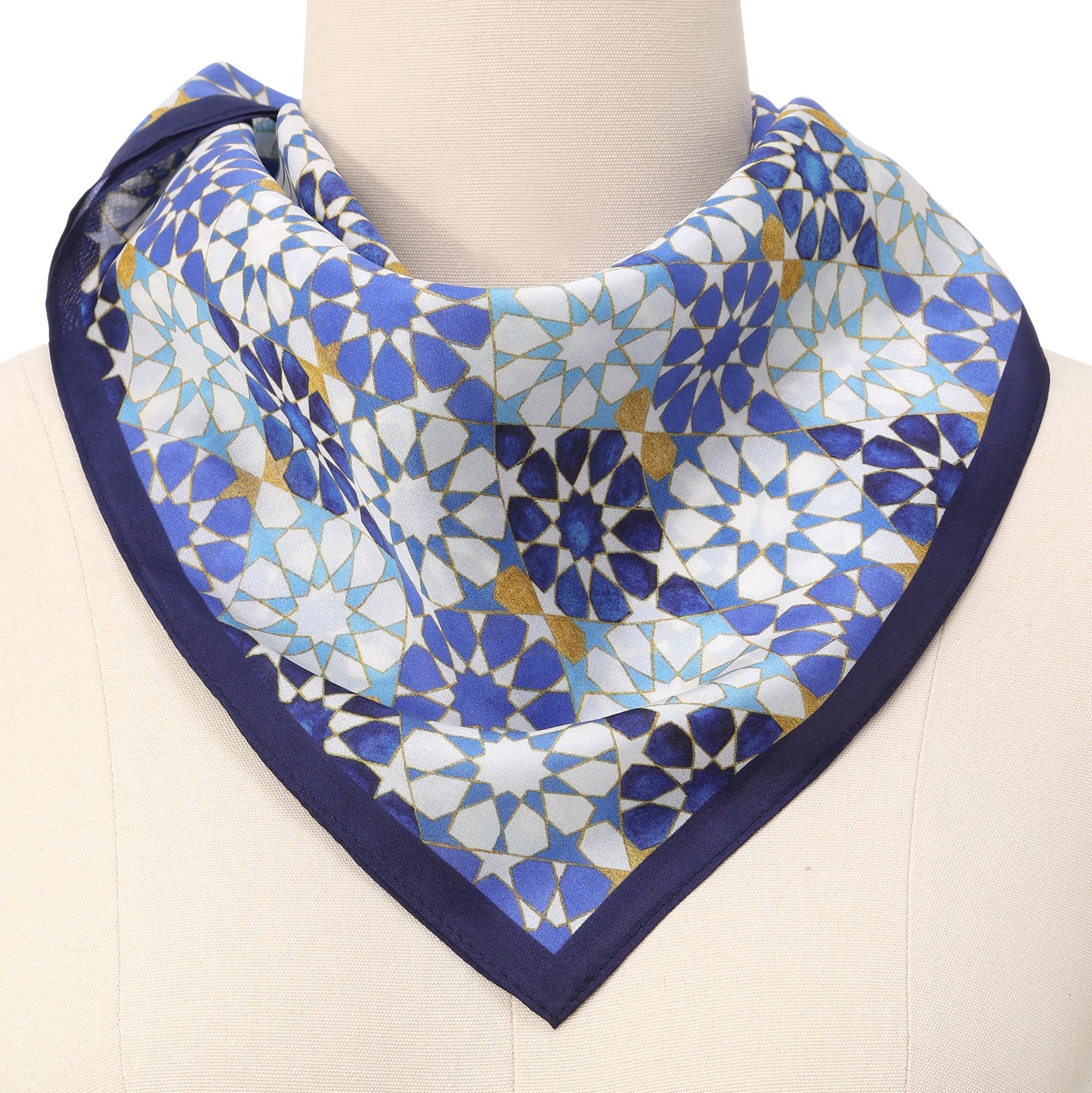 A mannequin wears the Blue Neck Scarf 100% Silk, featuring a geometric mosaic pattern in blue, white, and gold with a dark blue border. The scarf is neatly folded around the neck for a stylish look.
