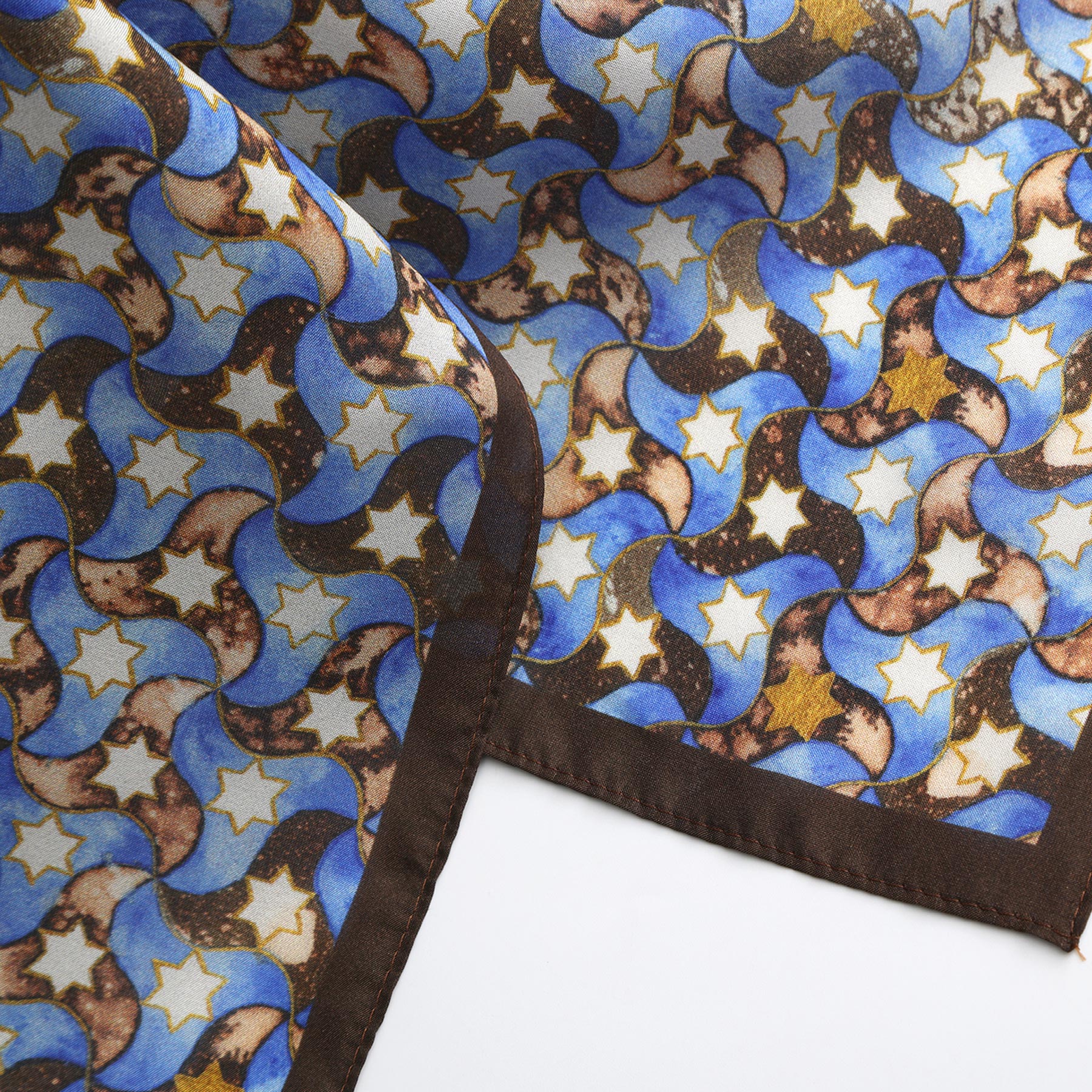 A close-up of the Silk Square Scarf for Women Brown, showcasing blue and brown crescent moons and yellow stars on silk with a dark brown stitched border. The scarf is slightly folded on a white surface.