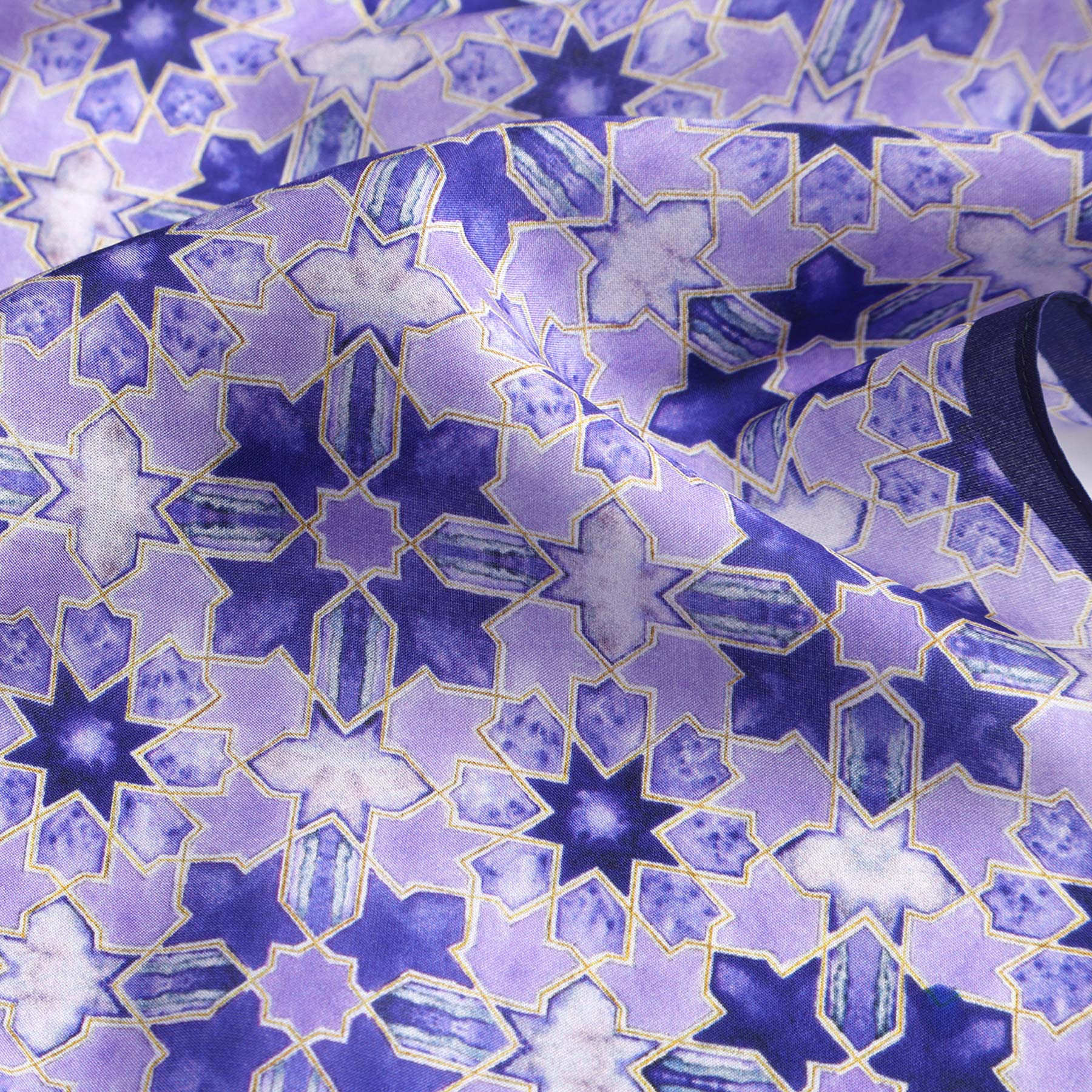 A close-up of the Square Silk Scarf for Women in purple features a geometric star pattern in blues, purples, and white with gold outlines. The silk material is slightly wrinkled and reflects light beautifully.