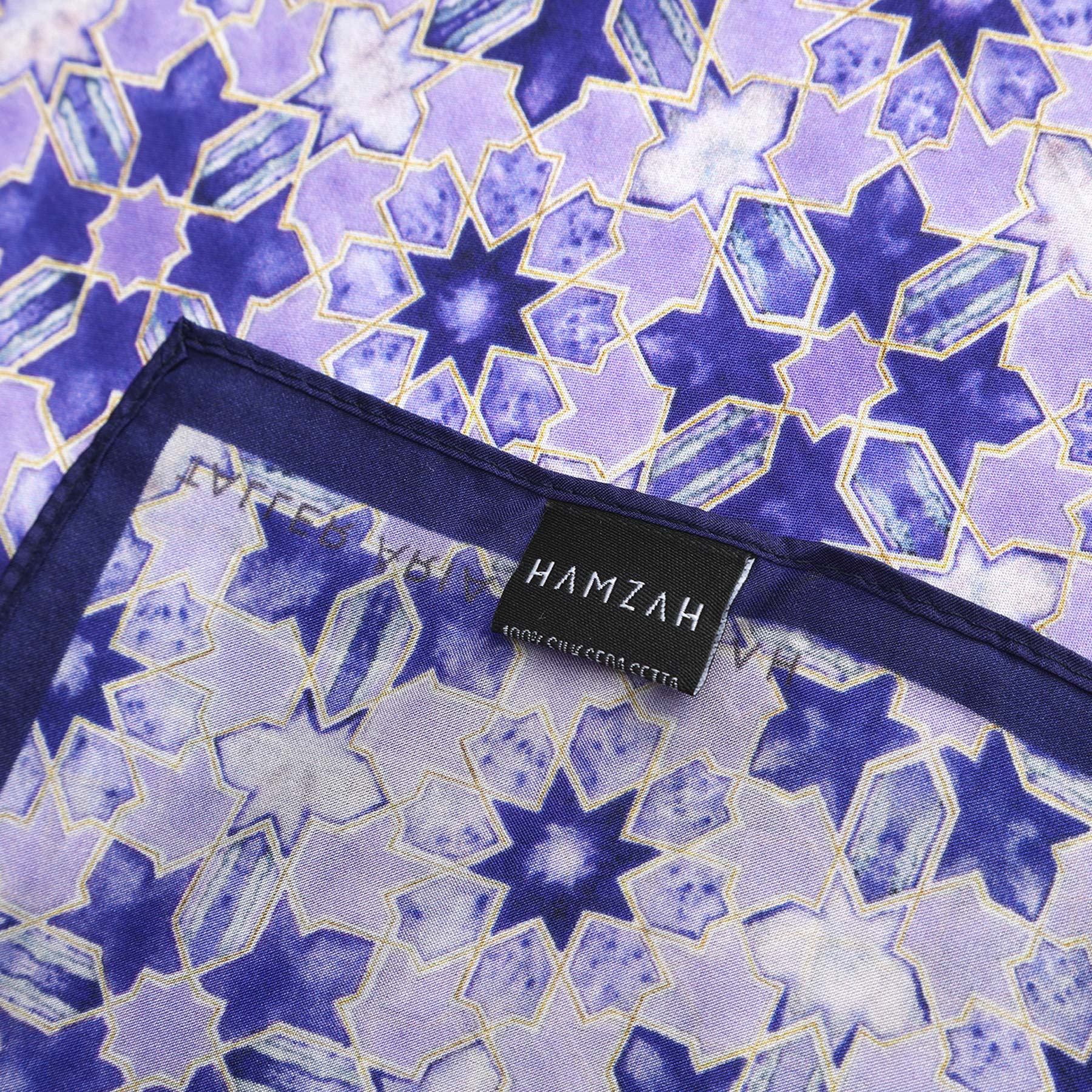 A close-up of the Square Silk Scarf for Women in purple, featuring geometric star patterns in blue and purple. A black label reading "HAMZAH 100% SUCCESS SETTA" is sewn onto the fabric.