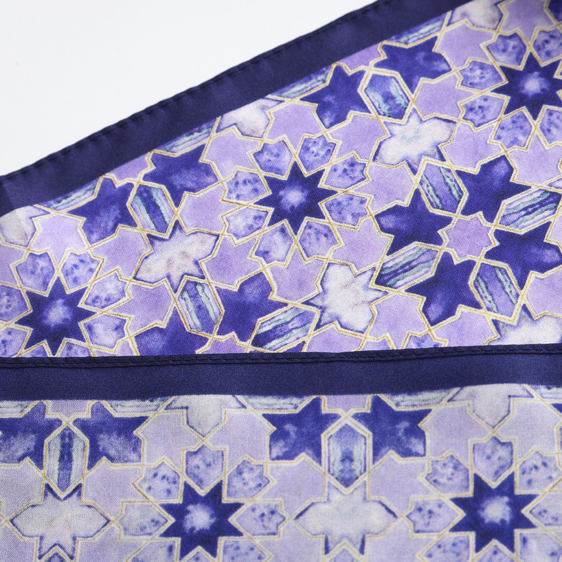 A close-up of the Square Silk Scarf for Women in purple showcases an intricate geometric pattern in purple, lavender, and blue with gold outlines. Interlocking star shapes fill the scarf, bordered by a solid dark blue edge.
