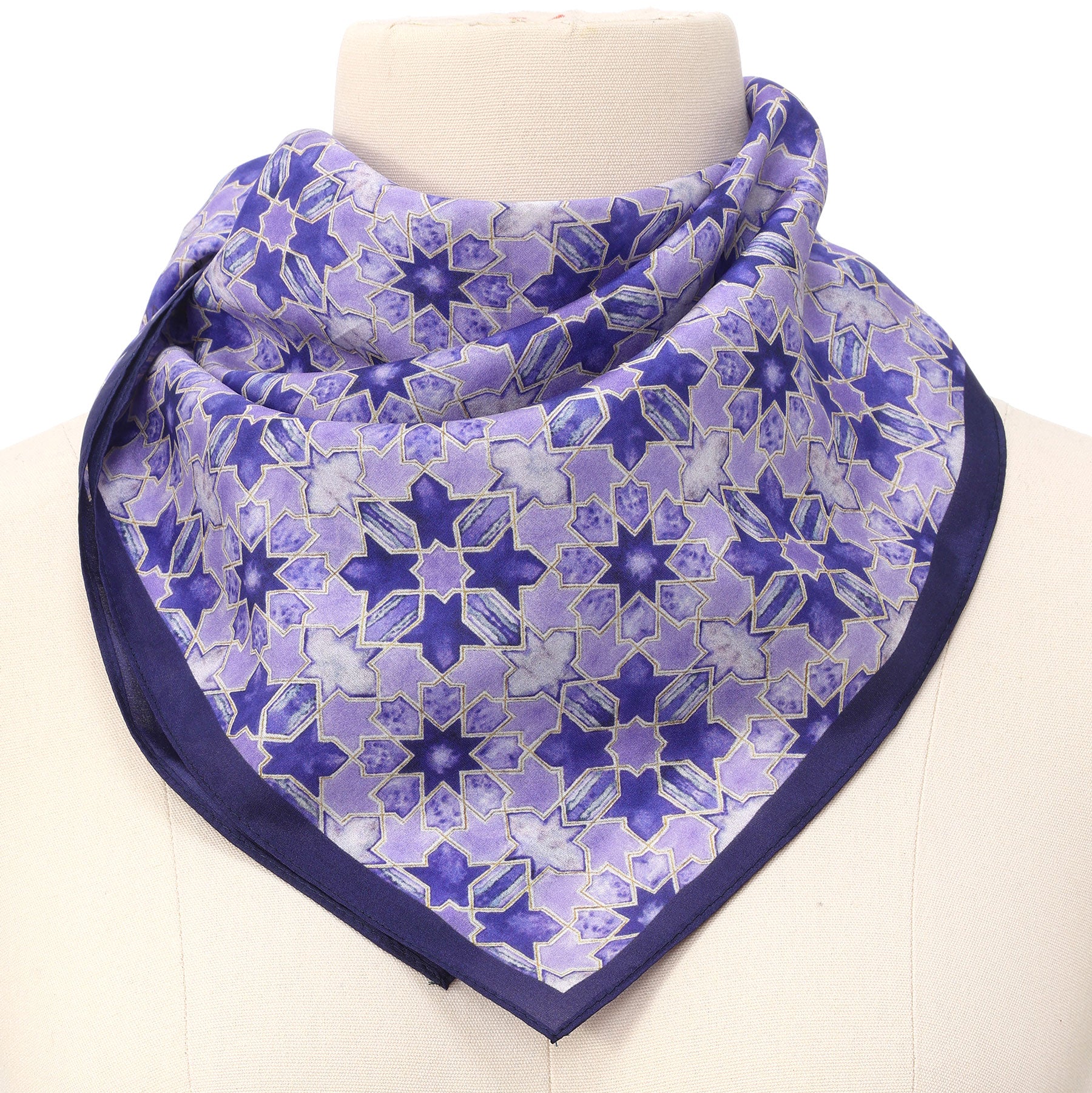 A mannequin displays the Square Silk Scarf for Women in purple, featuring a geometric star pattern and dark purple border, folded into a triangle and wrapped around the neck.