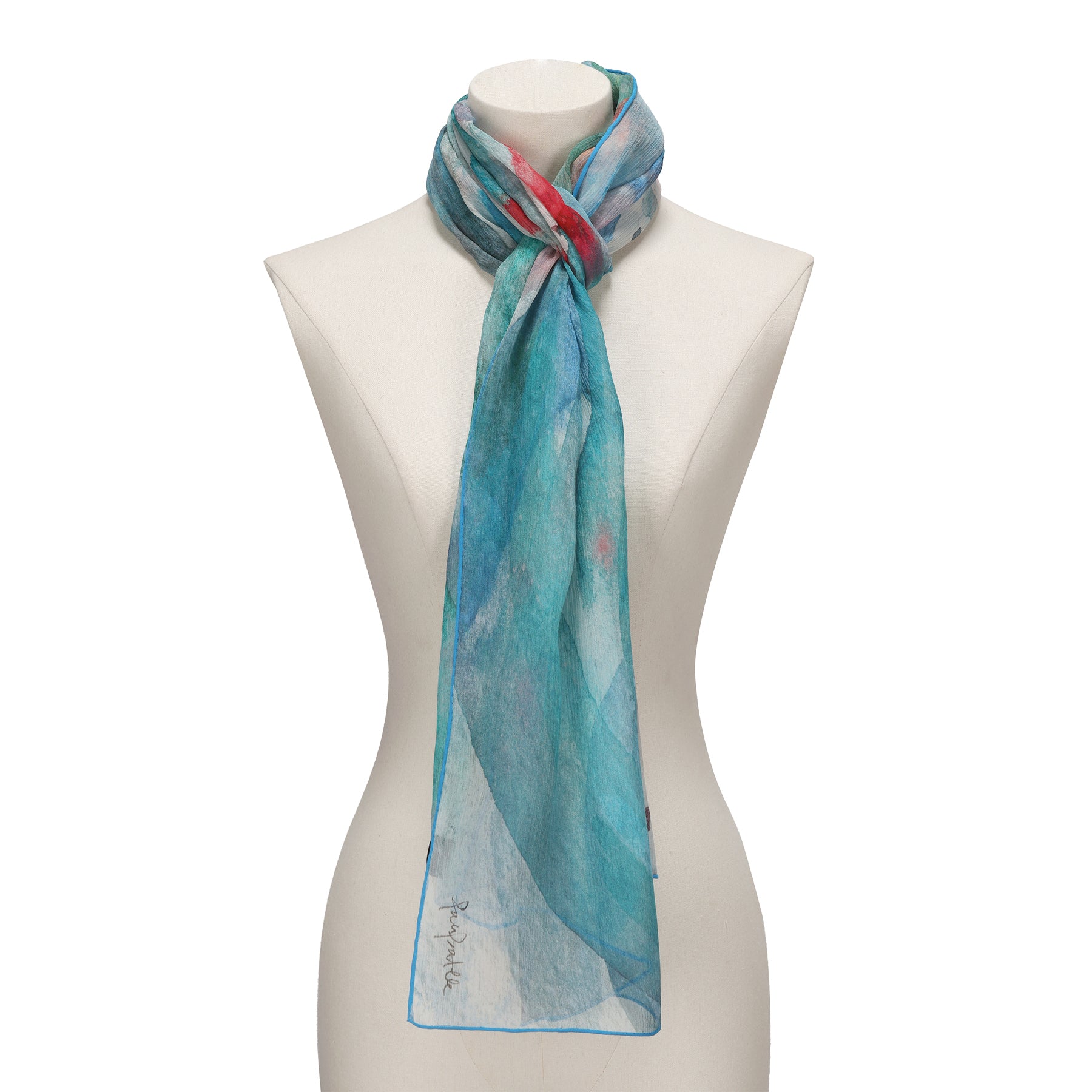 A mannequin showcases the Blue Silk Foulard Mediterráneos—an airy, slightly sheer muslin silk scarf in turquoise with red and white accents—loosely tied around the neck for an elegant Mediterranean touch.