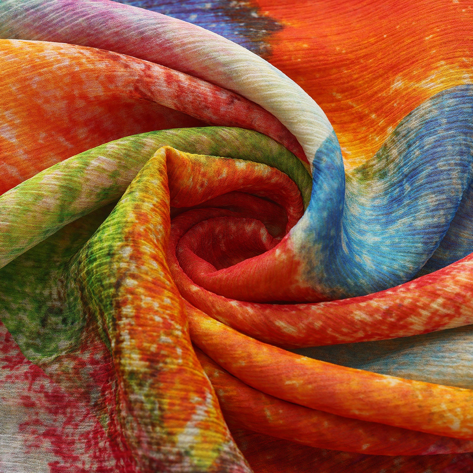A close-up of the Multicolor Silk Scarf Calidoscopic reveals vibrant swirls of orange, red, green, blue, and yellow in a dynamic tie-dye pattern, folded in a spiral shape that resembles wearable art.