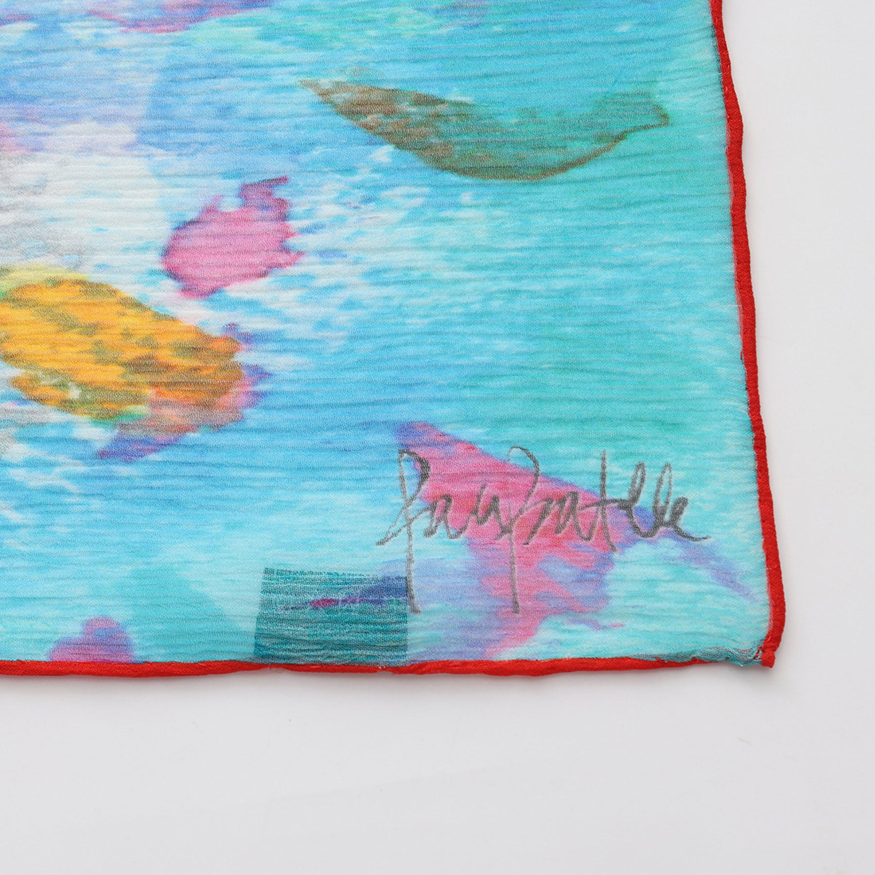 The Multicolor Silk Scarf Mar Rojo is a vibrant muslin silk scarf with abstract blue, pink, green, and yellow patterns bordered in red. It features a signature in the lower right corner, possibly reading Papoulatte.
