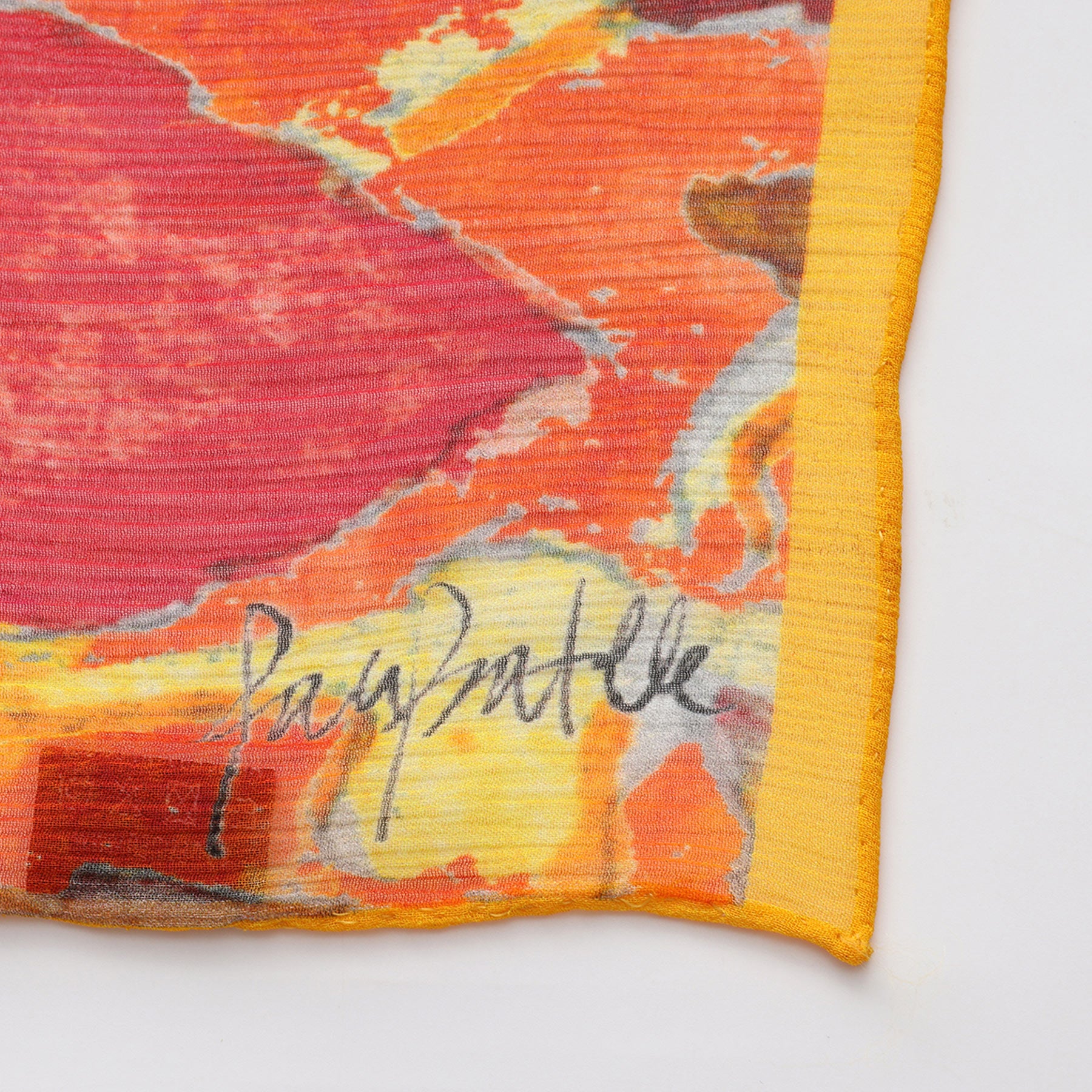 A close-up of the Multicolor Silk Scarf Demeter in muslin silk, showcasing a vibrant abstract pattern in red, orange, and yellow with a yellow hem and black-ink signature—perfect as wearable art.