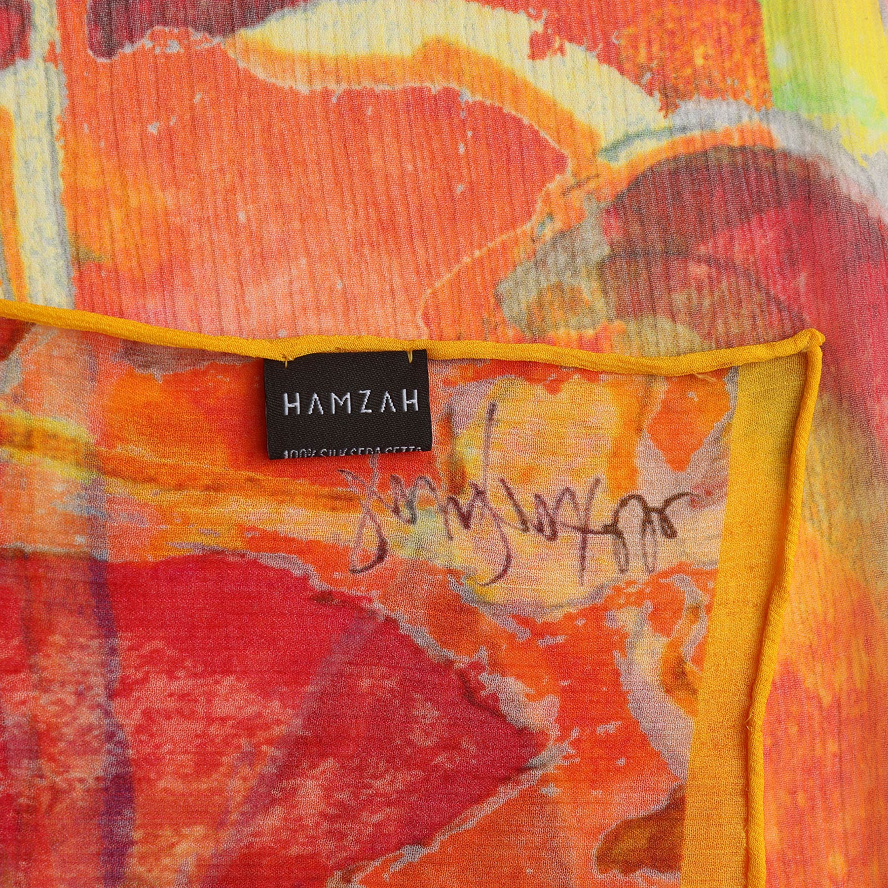 A close-up of the Multicolor Silk Scarf Demeter: a vibrant muslin silk accessory with red, yellow, and orange abstract designs, adorned with a black HAMZAH label and a handwritten signature near the fabric’s edge.