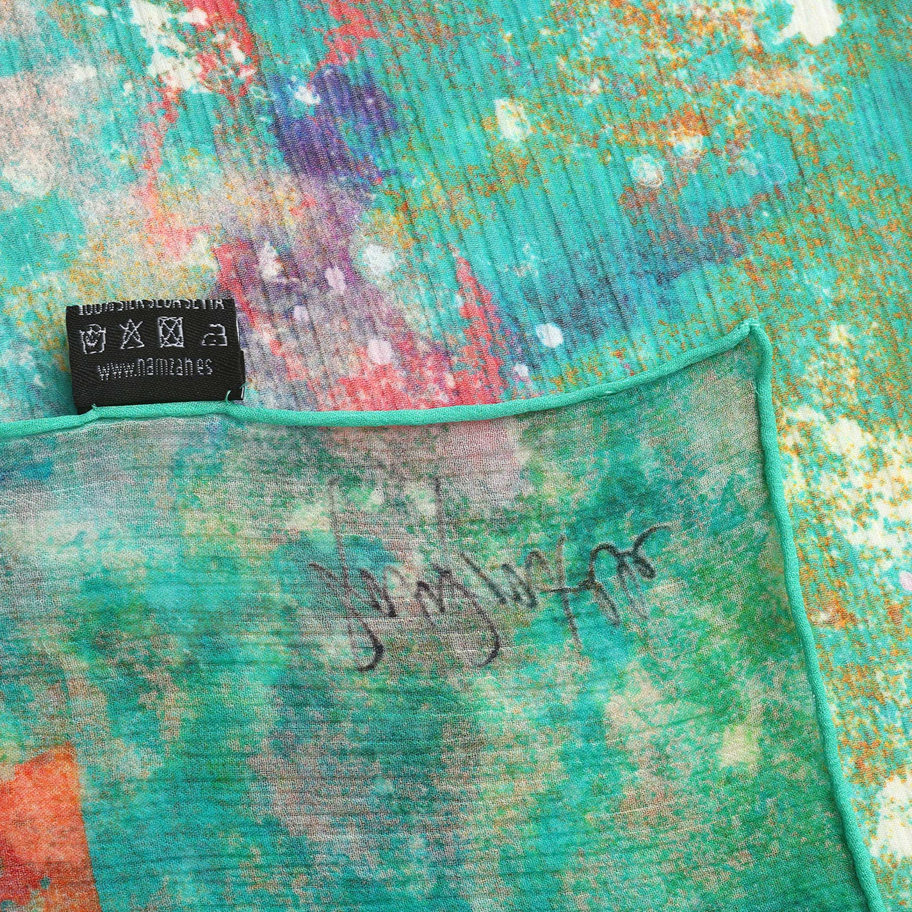 A close-up of the Natural Silk Scarf Mundos Ocultos showcases its colorful abstract pattern in turquoise, yellow, and red tones, with a black clothing tag and handwritten signature on the edge—a true piece of wearable art.