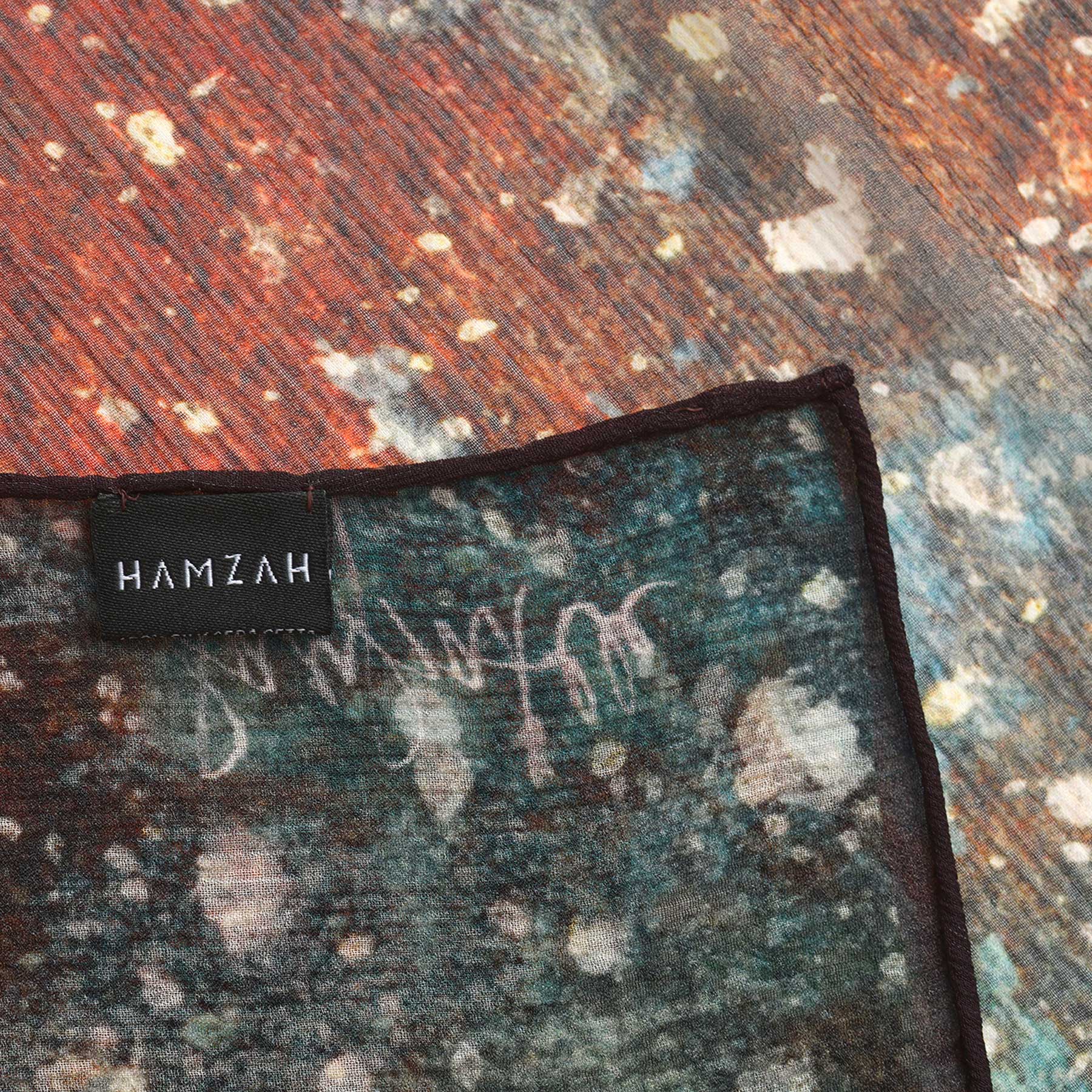 A close-up of the Multicolor Silk Scarf Polvo Cósmico shows its abstract, vibrant pattern. A black HAMZAH label accents the muslin silk, while the background reflects the scarf’s cosmic-inspired design, celebrating this unique wearable artwork.