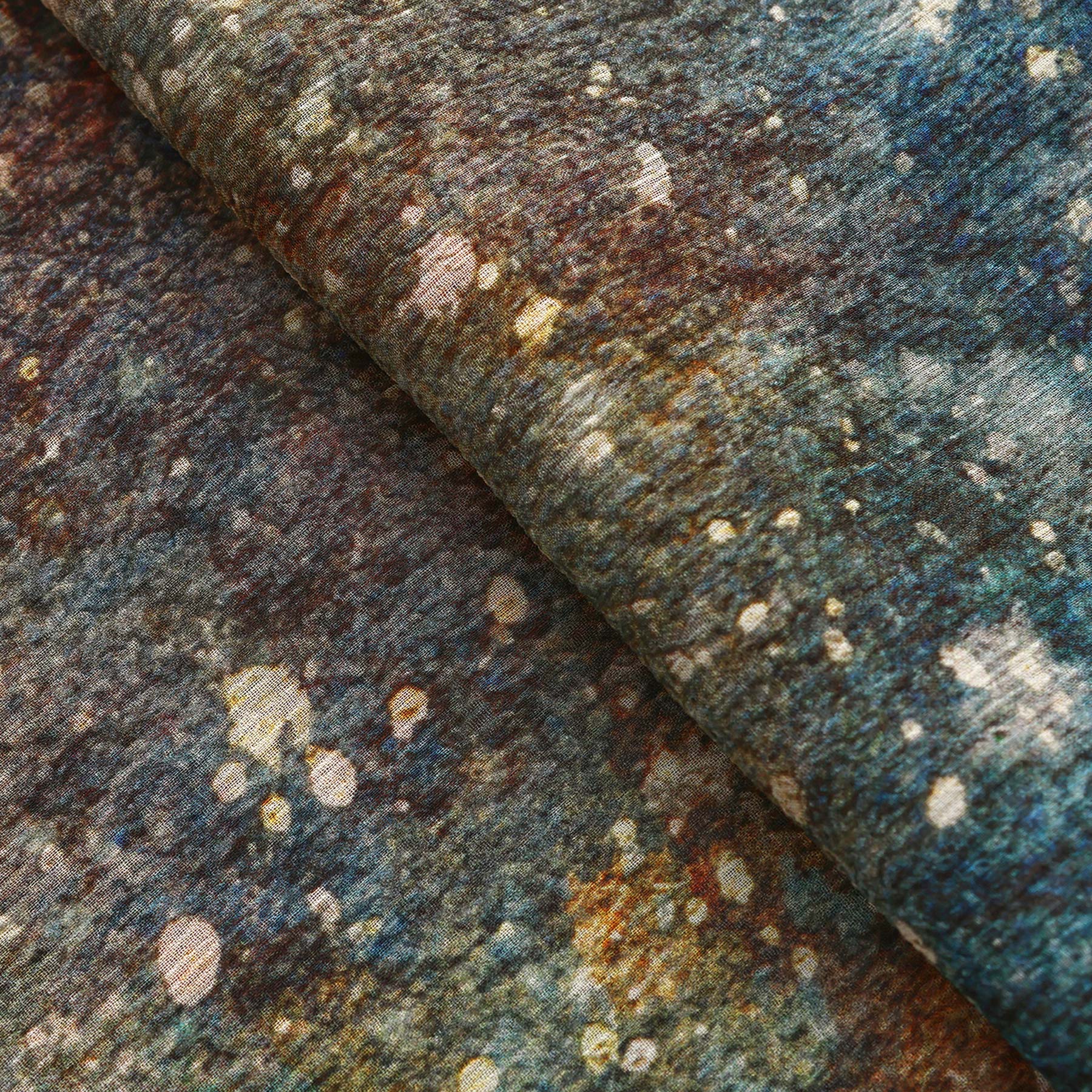 A close-up of the Multicolor Silk Scarf Polvo Cósmico shows a muslin silk fabric with blue, brown, yellow, and white speckles in an abstract pattern—like splattered paint or a starry sky—perfect as wearable art.