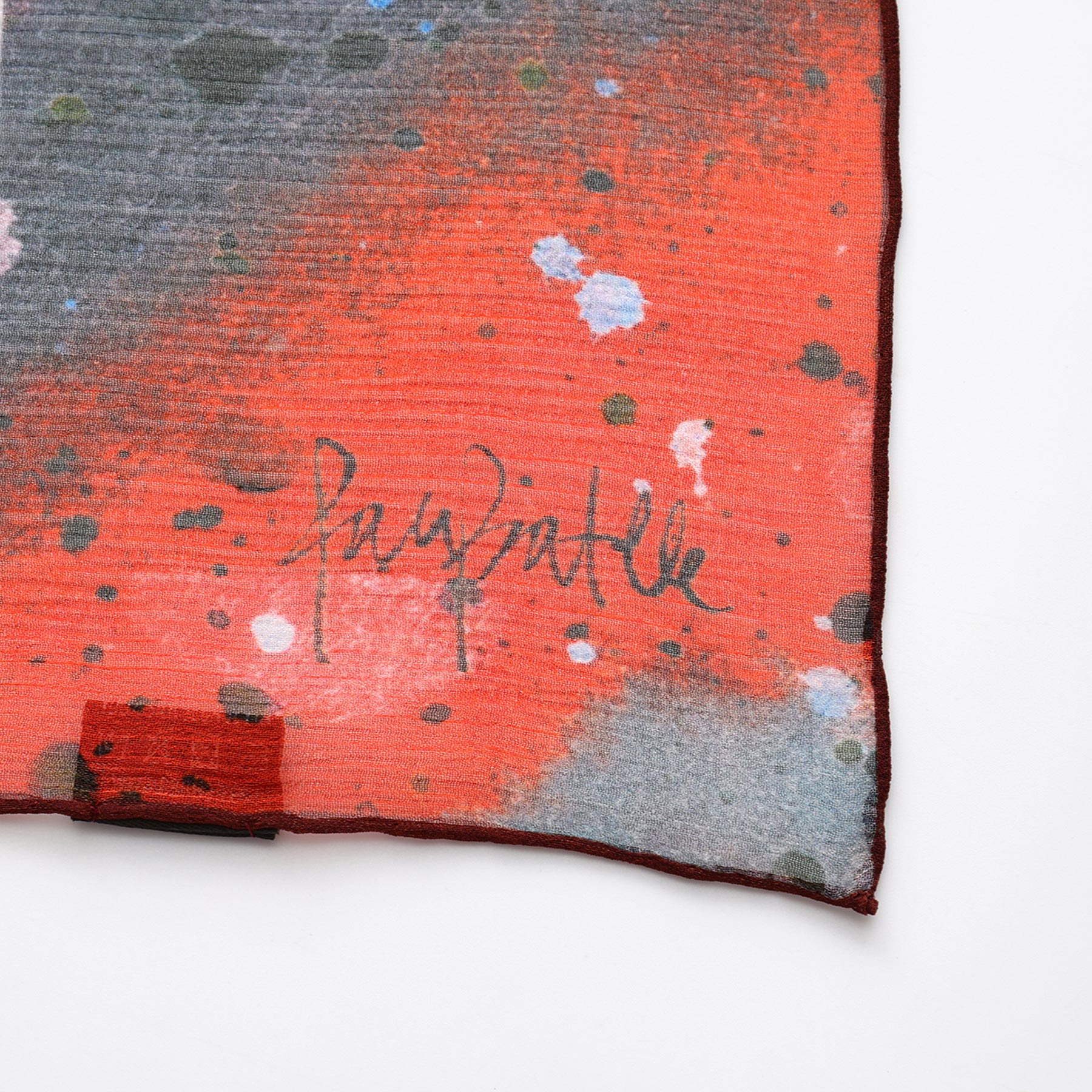 A close-up of the Multicolor Silk Scarf Cangrejos shows paint splatter patterns and a distinctive signature near the dark, slightly curled edge, highlighting this wearable artworks unique style.