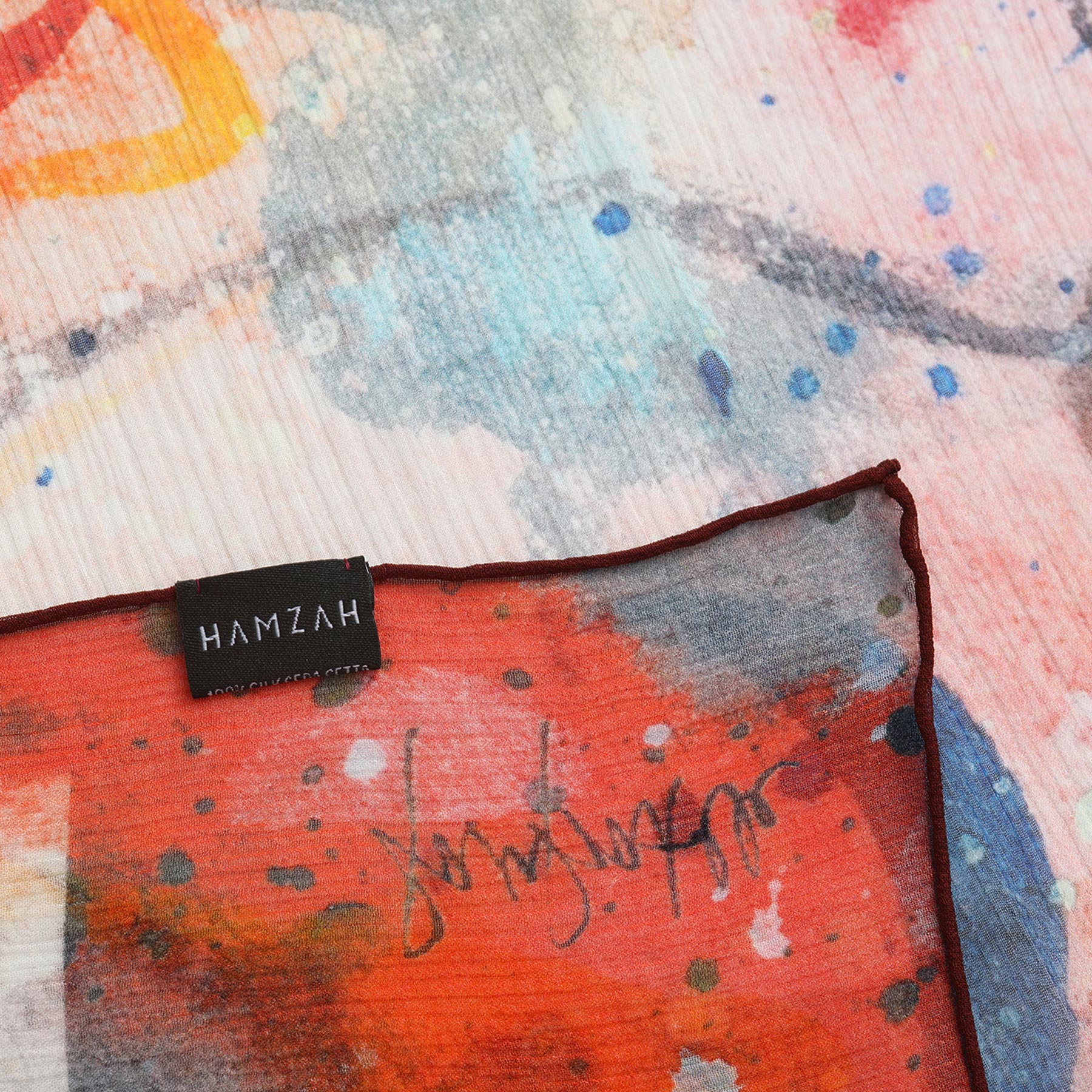 A close-up of the Multicolor Silk Scarf Cangrejos shows vibrant abstract patterns in red, blue, and orange, with a black HAMZAH label on the edge—embodying luxurious Mediterranean style in wearable art.