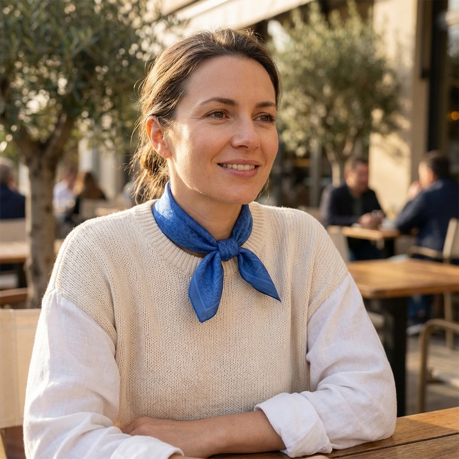 A woman with brown hair tied back, wearing a cream sweater and the Blue Square Silk Scarf Dalila v1, sits smiling at an outdoor café table. Trees and people can be seen in the background.
