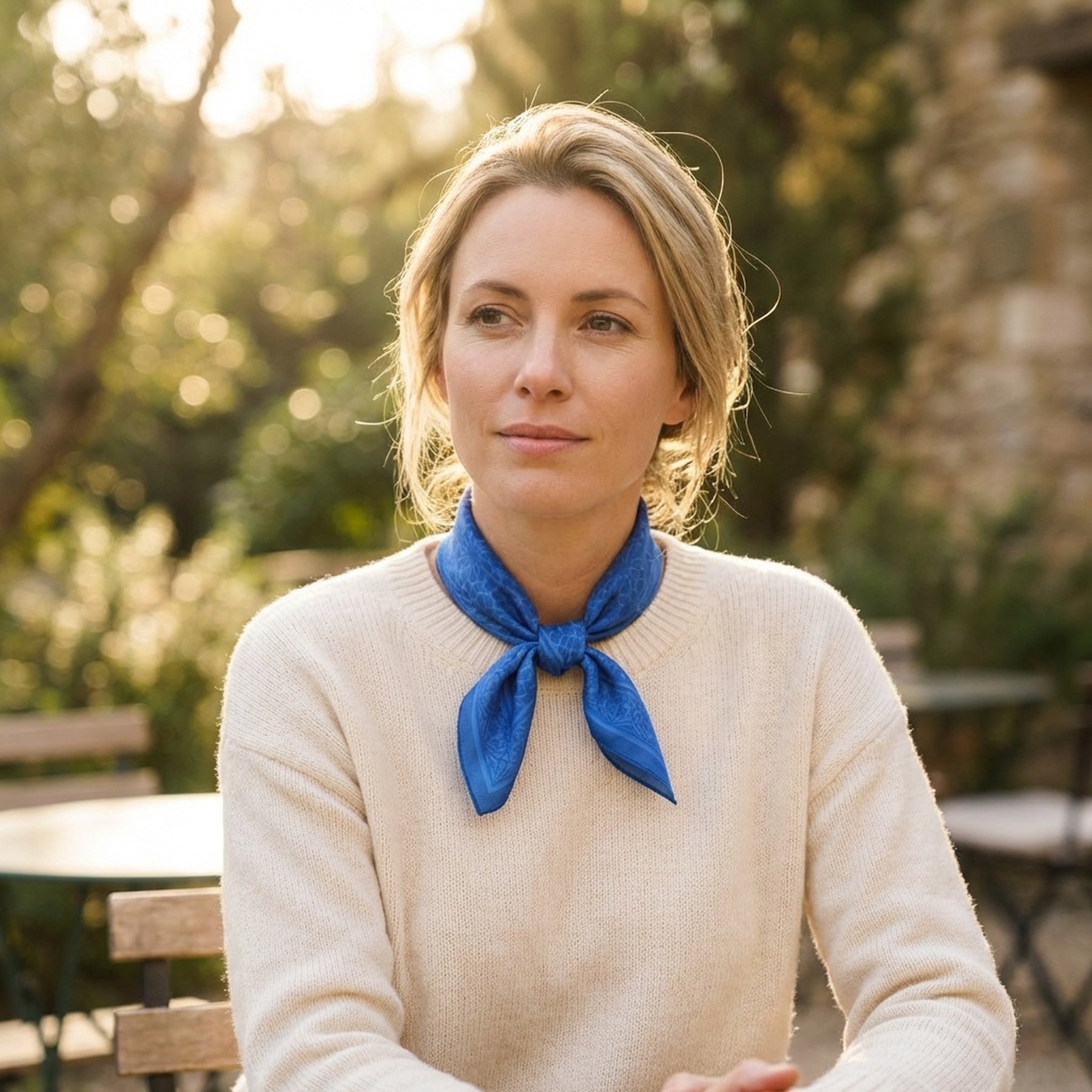 A woman with blonde hair, in a cream sweater, wears the Blue Square Silk Scarf Dalila v1 at her neck while sitting outdoors amid sunlit garden greenery. An Alhambra Palace-inspired Islamic art print is placed beside her.