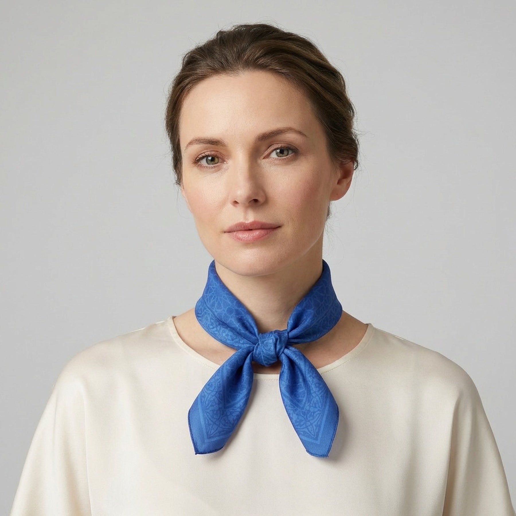 A woman with light skin and brown hair pulled back stands against a light gray background, wearing a cream blouse and the Blue Square Silk Scarf Dalila v1, featuring a pattern inspired by Islamic art prints.