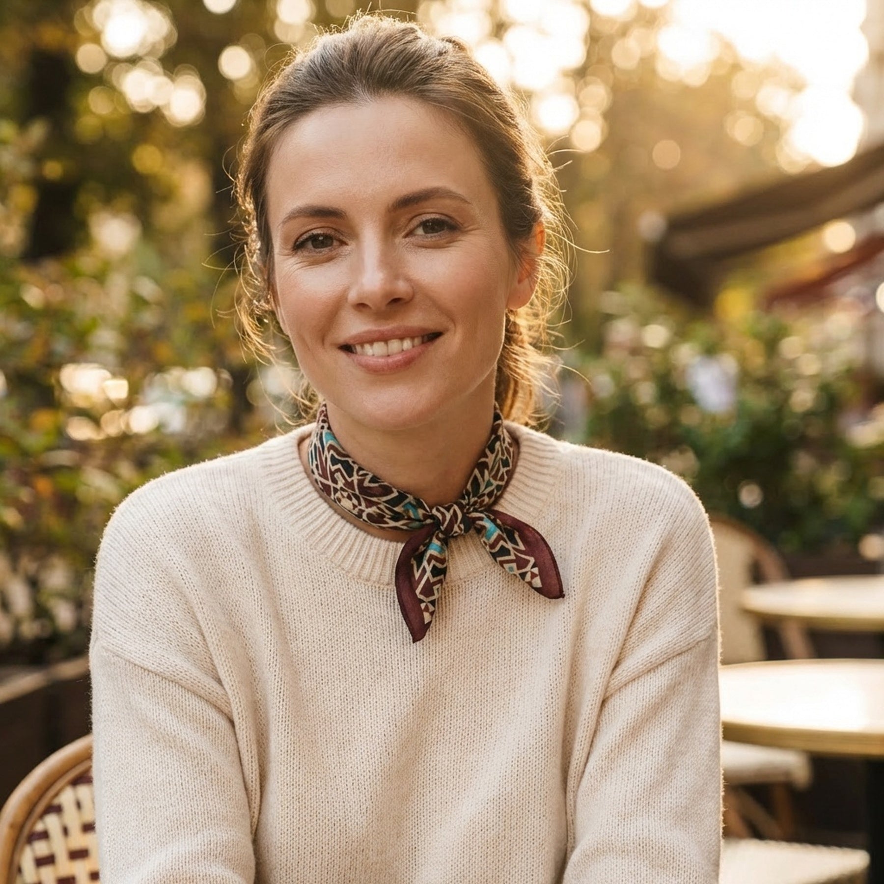 A woman with light brown hair tied back, in a cream sweater and the Brown Silk Neckerchief, sits smiling at an outdoor café. Sunlight highlights greenery and empty chairs in the background.