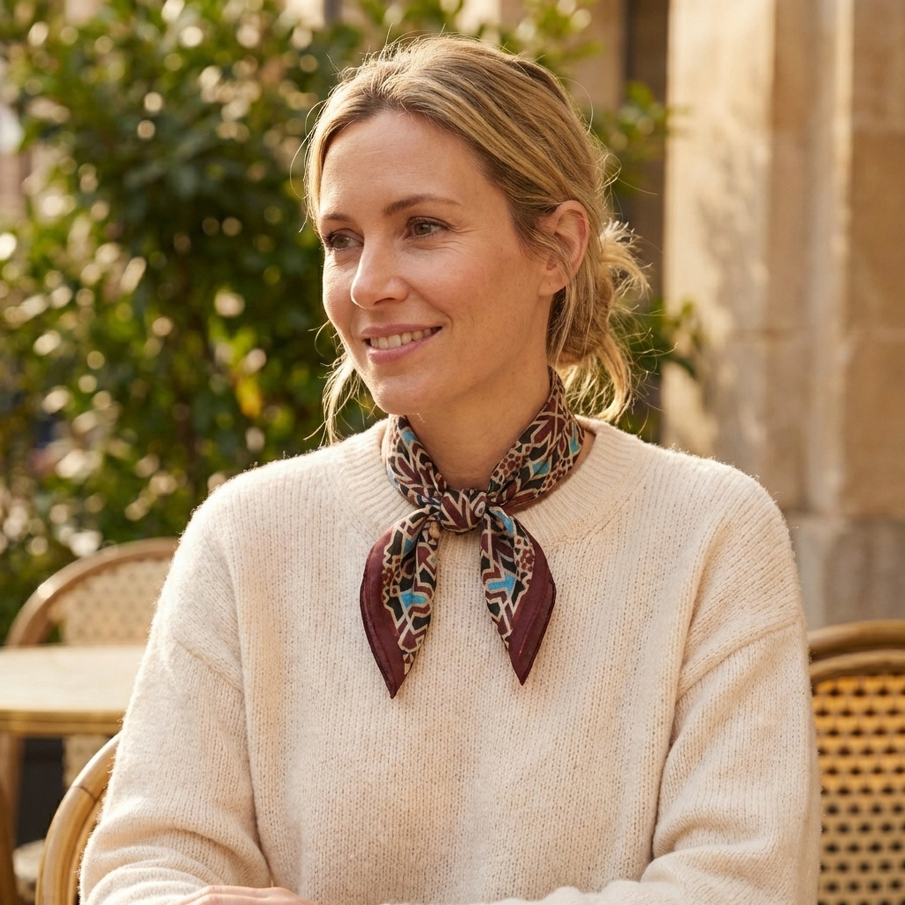 A smiling woman with blonde hair tied back sits at an outdoor café table, wearing a cream sweater and the Brown Silk Neckerchief, which features an Islamic art print. Green plants and stone columns are in the background.