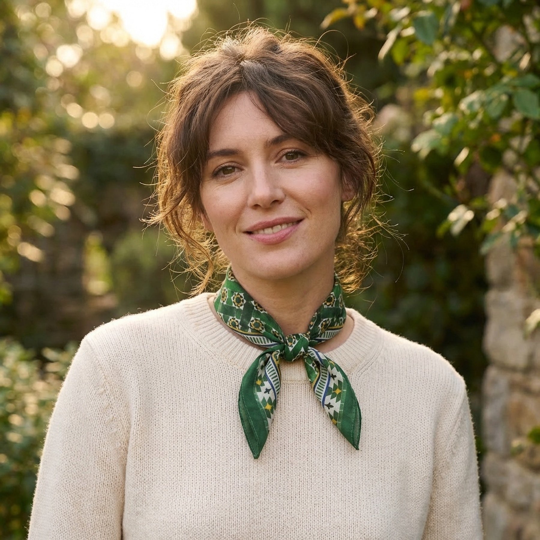 A woman with wavy brown hair smiles gently outdoors, wearing a cream sweater and the Green Square Scarf Silk tied around her neck, as sunlight filters through greenery in the background.