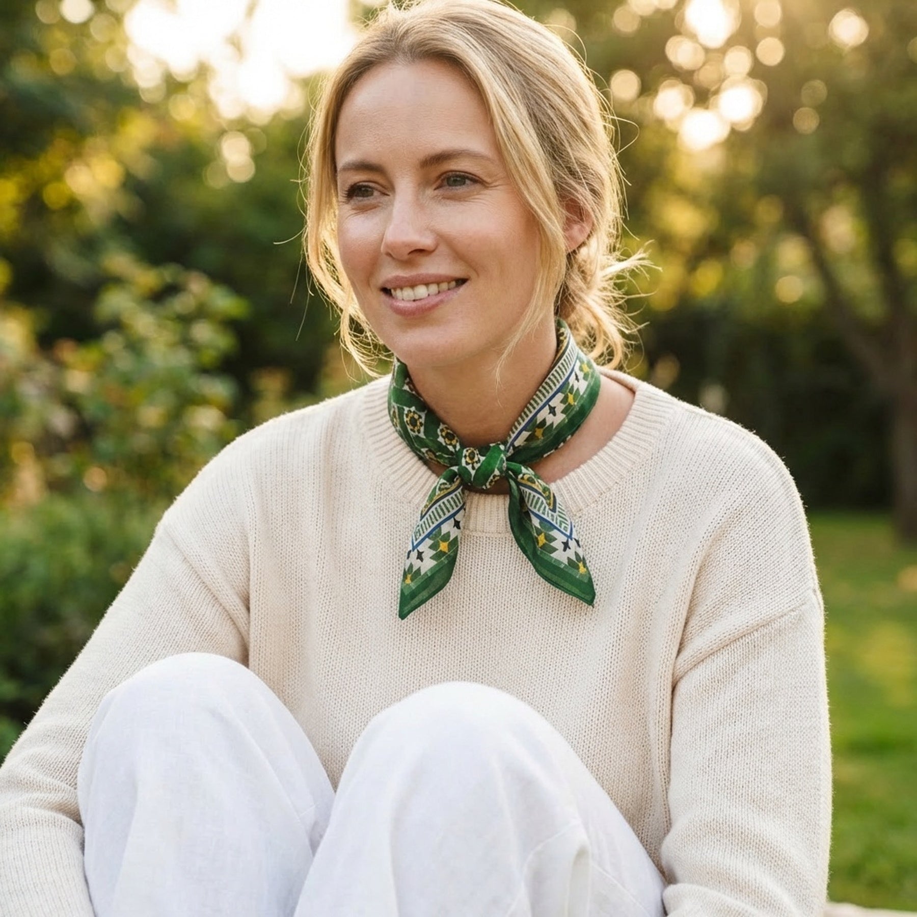 A smiling woman sits outdoors on grass, wearing a cream sweater, white pants, and the Green Square Scarf Silk around her neck as sunlight filters through trees in the background.