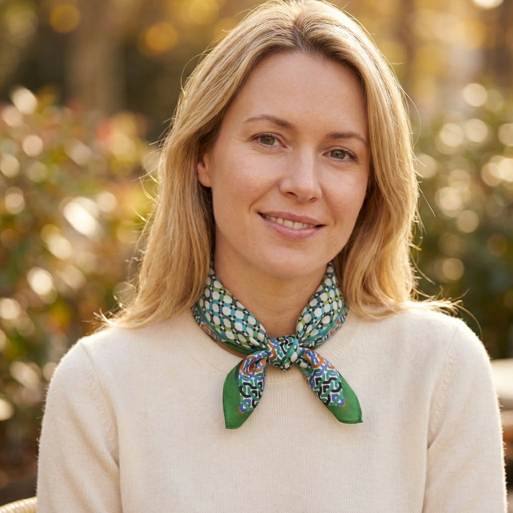 A blonde woman in a cream sweater smiles softly outdoors, wearing the Coloful Neckerchief 100% Silk. The blurred background features greenery and warm light.