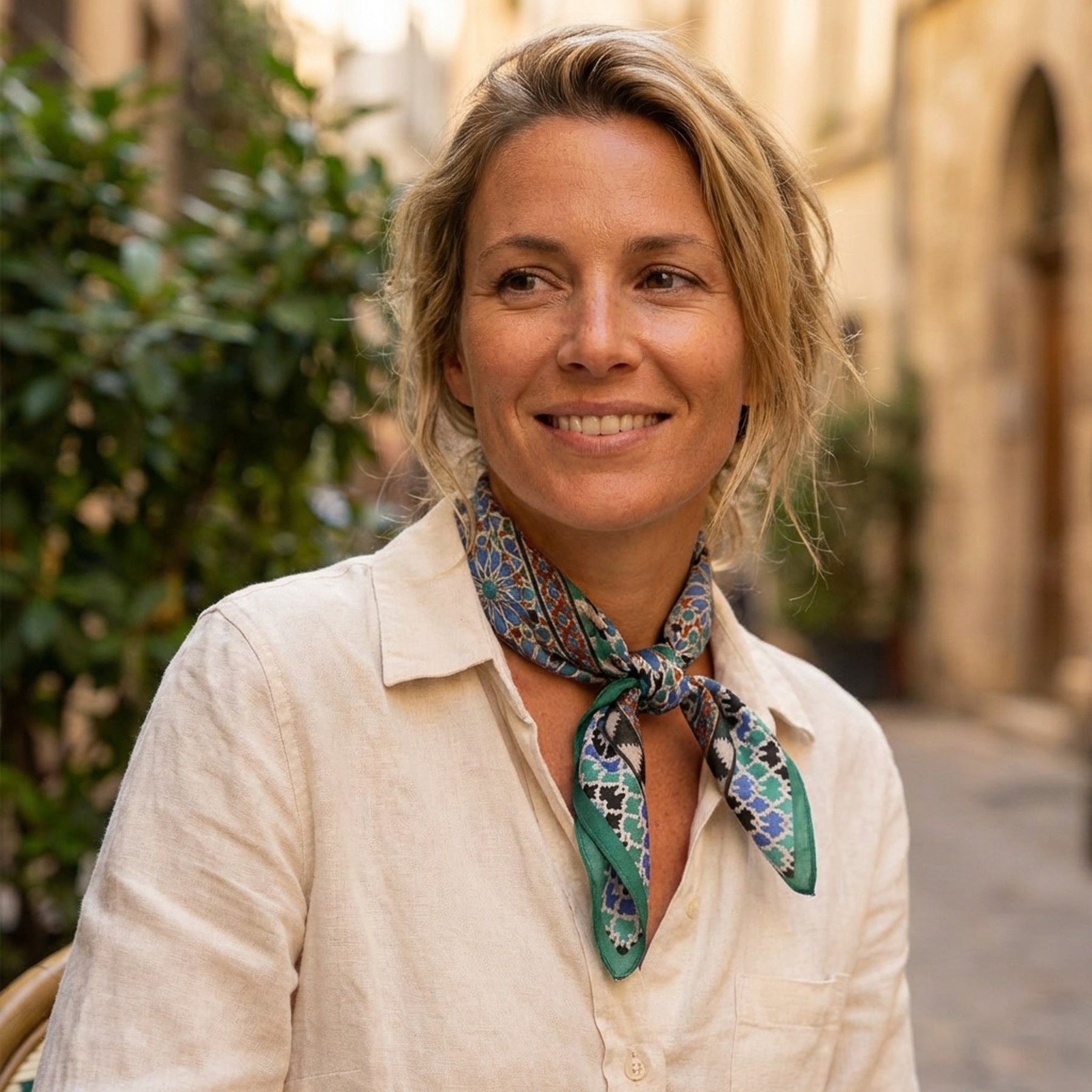 A blonde woman in a light beige shirt smiles outdoors on a sunny day, wearing the Alhambra Art Silk Square Scarf around her neck, with blurred buildings and greenery in the background.