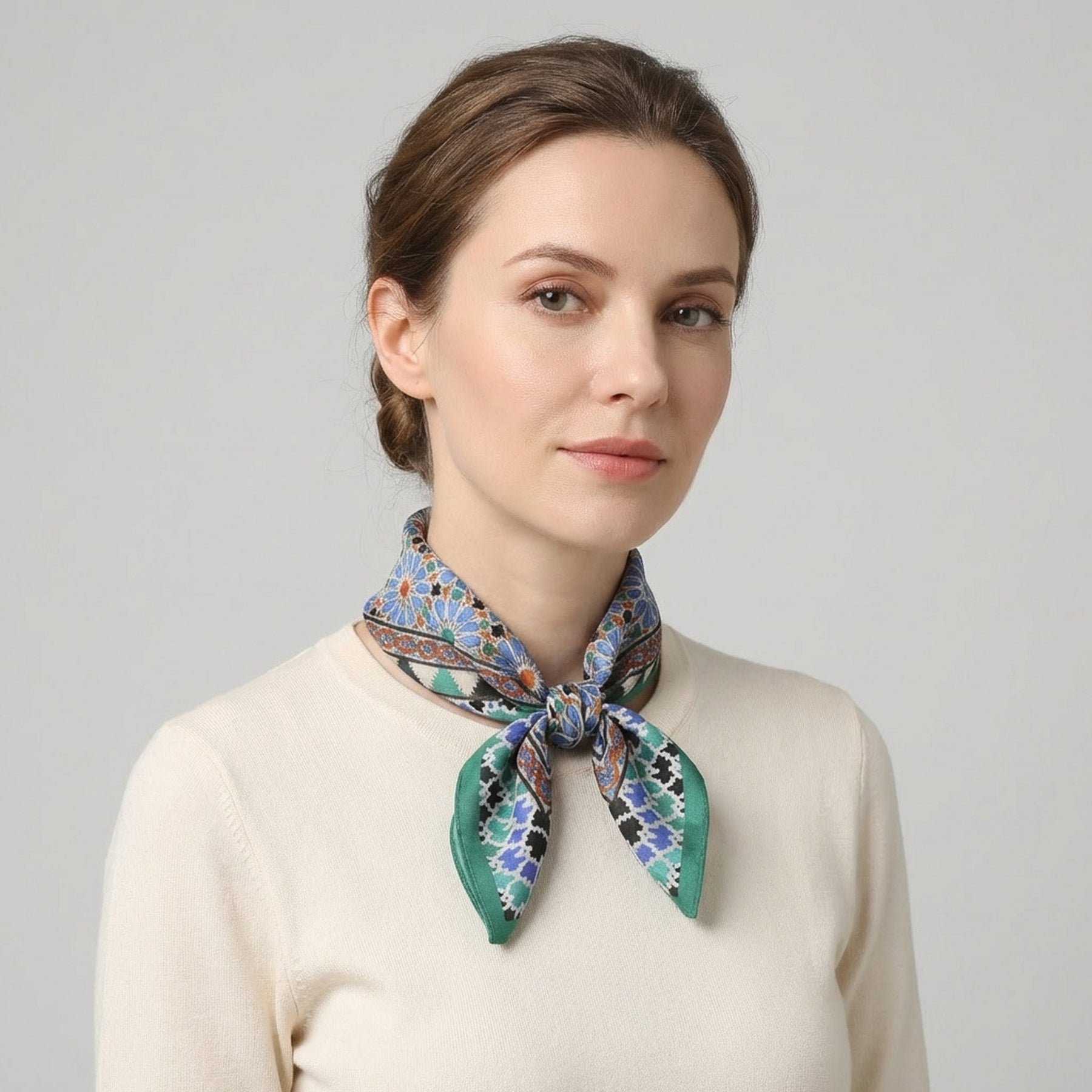 A woman with light brown hair in a low bun wears a cream sweater and the Alhambra Art Silk Square Scarf, featuring blue, green, and brown Islamic art-inspired accents, standing against a plain light gray background.