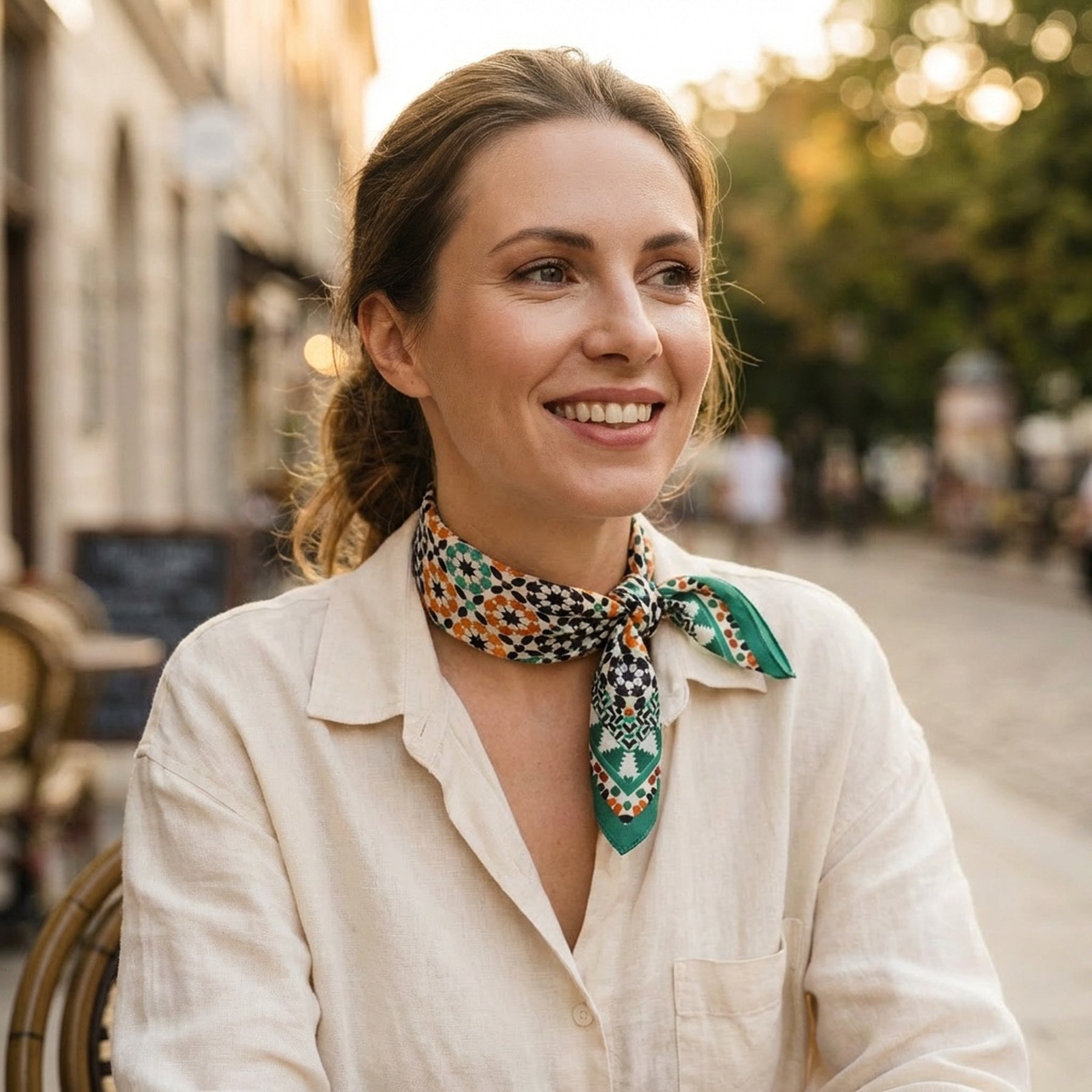 A woman with light brown hair tied back smiles at an outdoor cafe, wearing a cream shirt and the Green Square Silk Scarf Sabika, whose elegant drape reflects Spanish craftsmanship. Trees and blurred buildings are visible in the background.