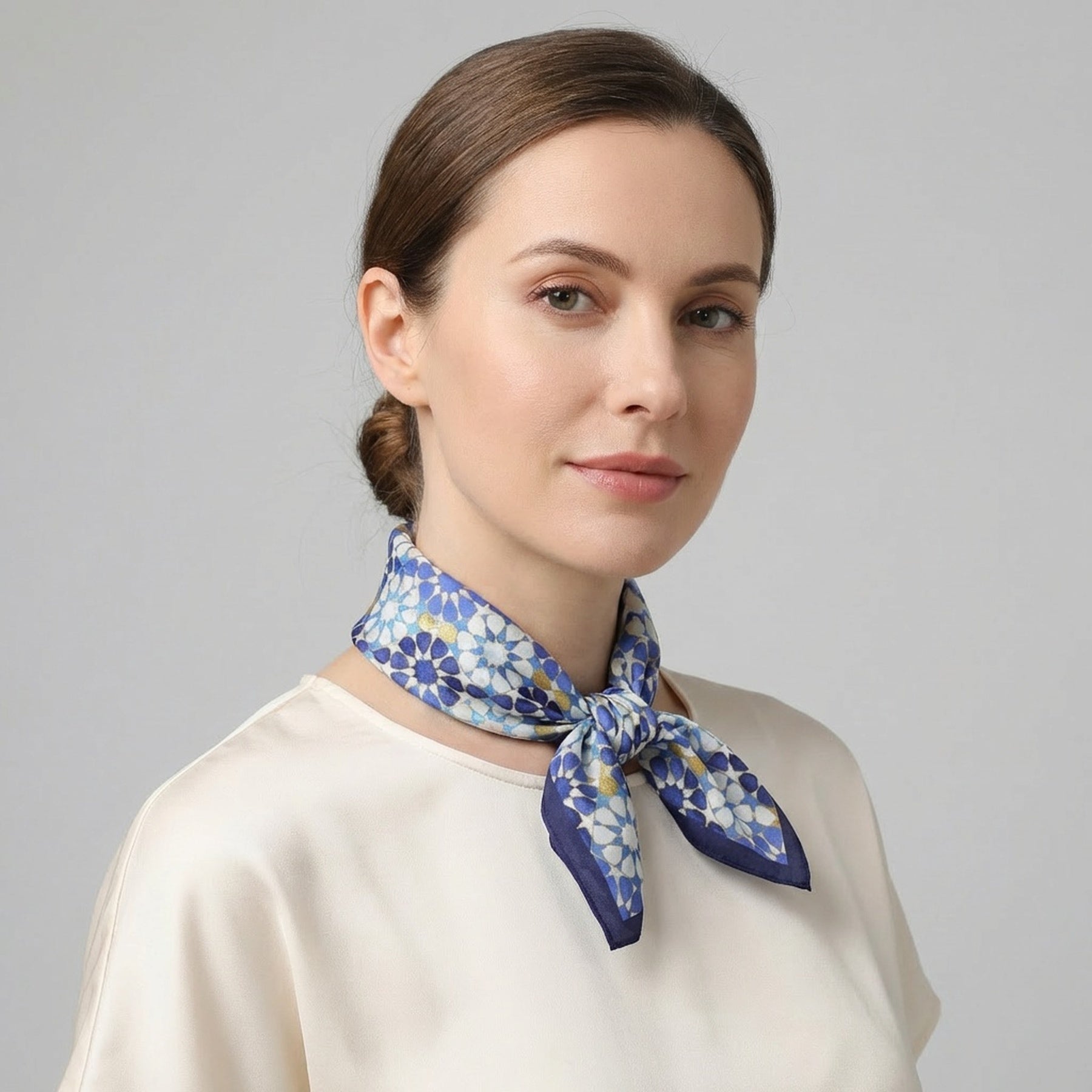 A woman with fair skin and brown hair tied back wears a cream blouse and the Blue Neck Scarf 100% Silk, neatly tied around her neck against a plain light gray background.