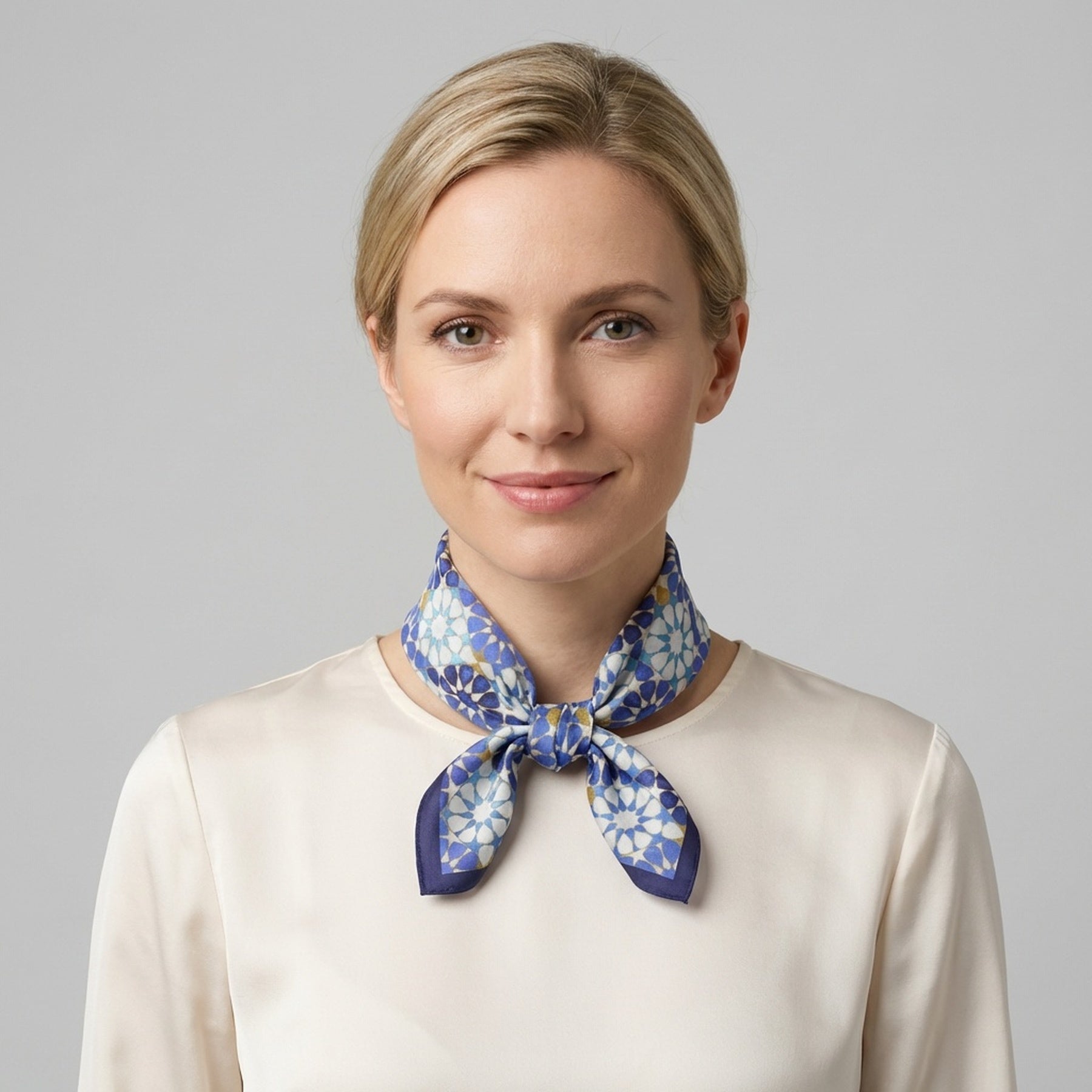 A woman with blonde hair in a low bun smiles gently against a plain light gray background, wearing a cream blouse and the Blue Neck Scarf 100% Silk elegantly tied around her neck.