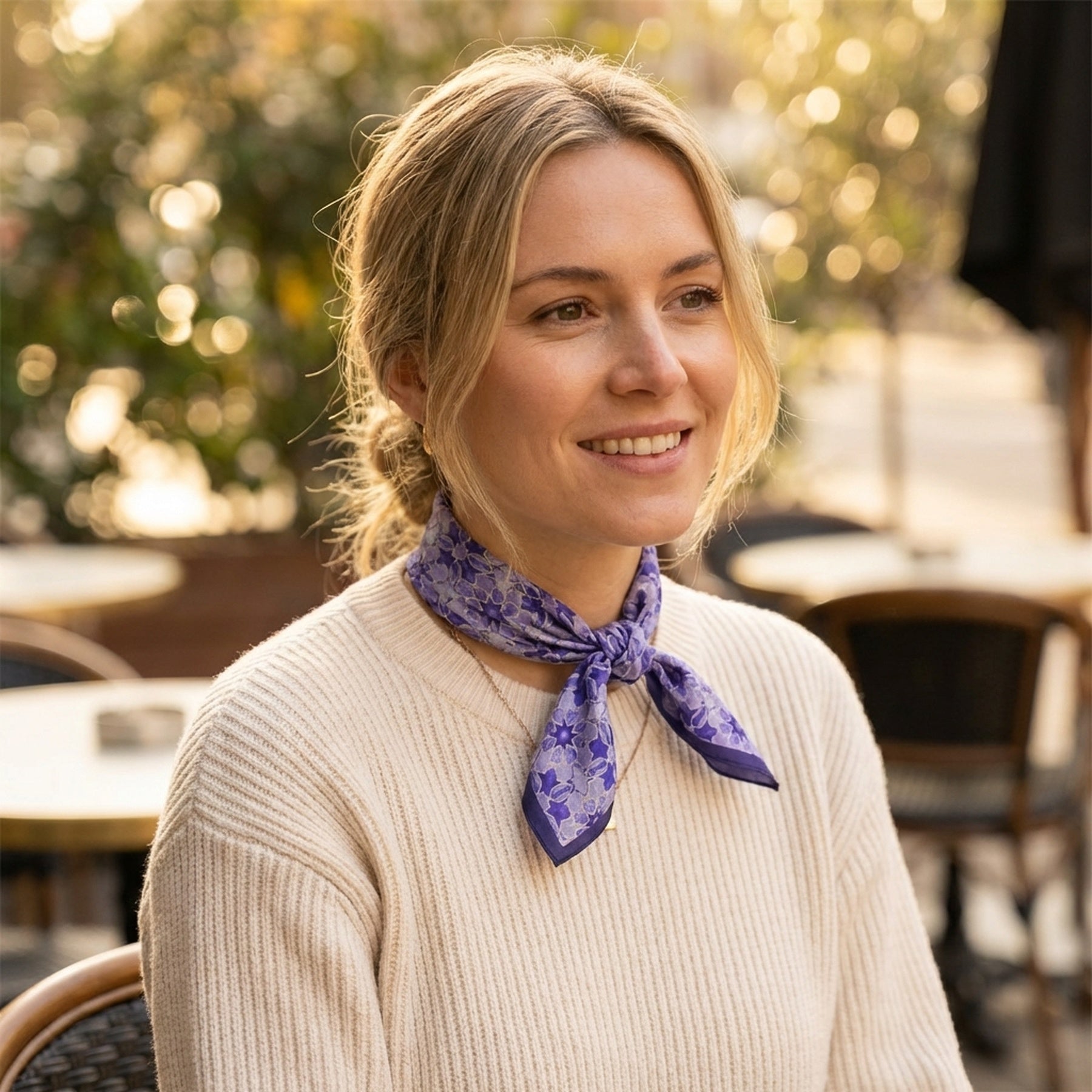 A smiling blonde woman with her hair tied back wears a cream sweater and the Square Silk Scarf for Women Purple around her neck while sitting at an outdoor café with blurred chairs and greenery in the background.