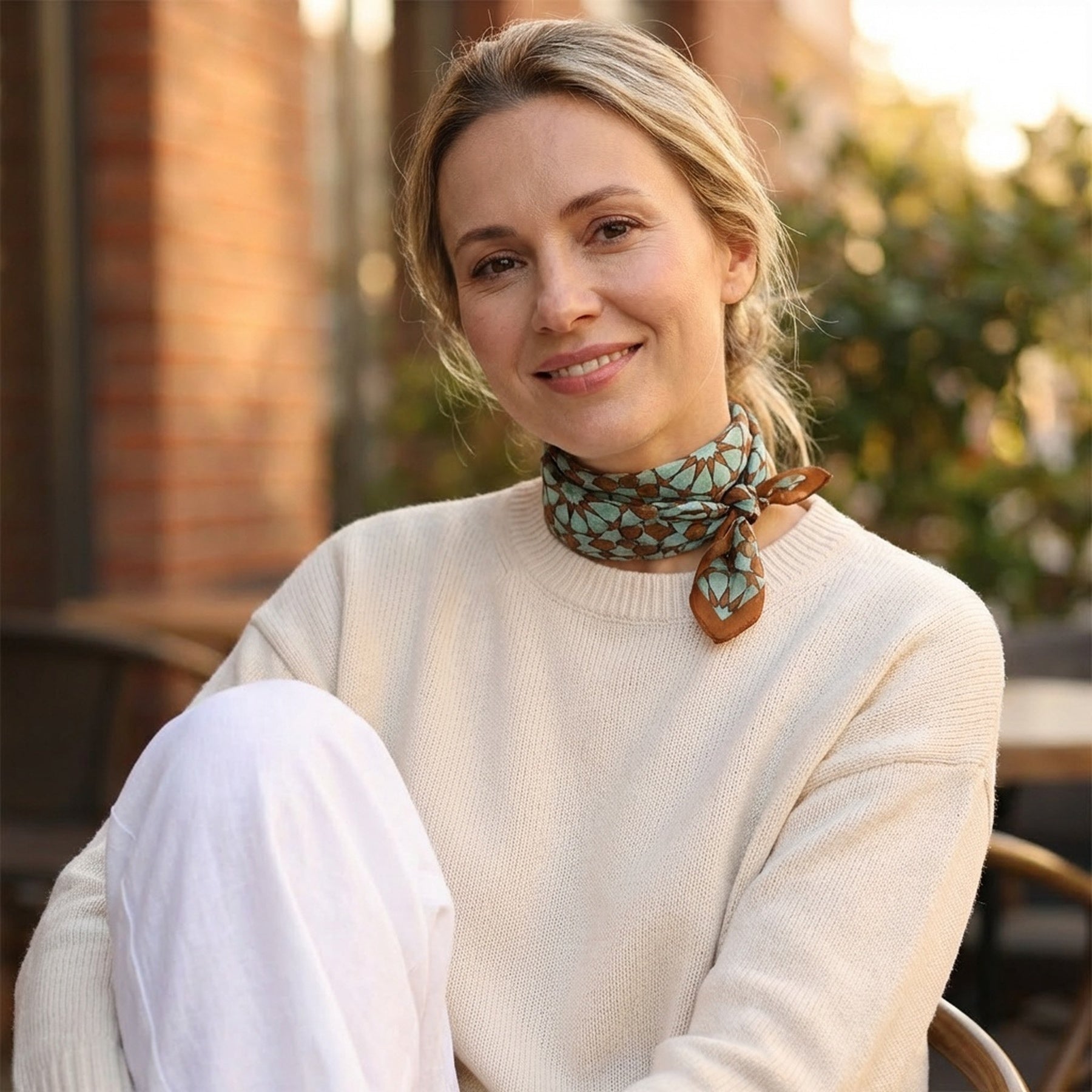 A smiling woman with blonde hair, wearing a cream sweater and the Brown Silk Neckerchief for Women, sits outdoors in a relaxed pose. Green plants and a brick wall are visible in the blurred background.