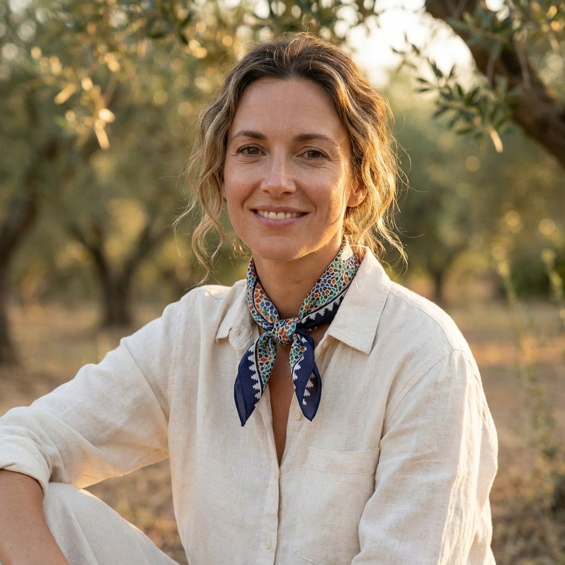 A woman with wavy hair, in a light linen shirt and the Square Scarf Blue Madiya inspired by Islamic art, sits outdoors among olive trees, smiling softly in warm sunlight.