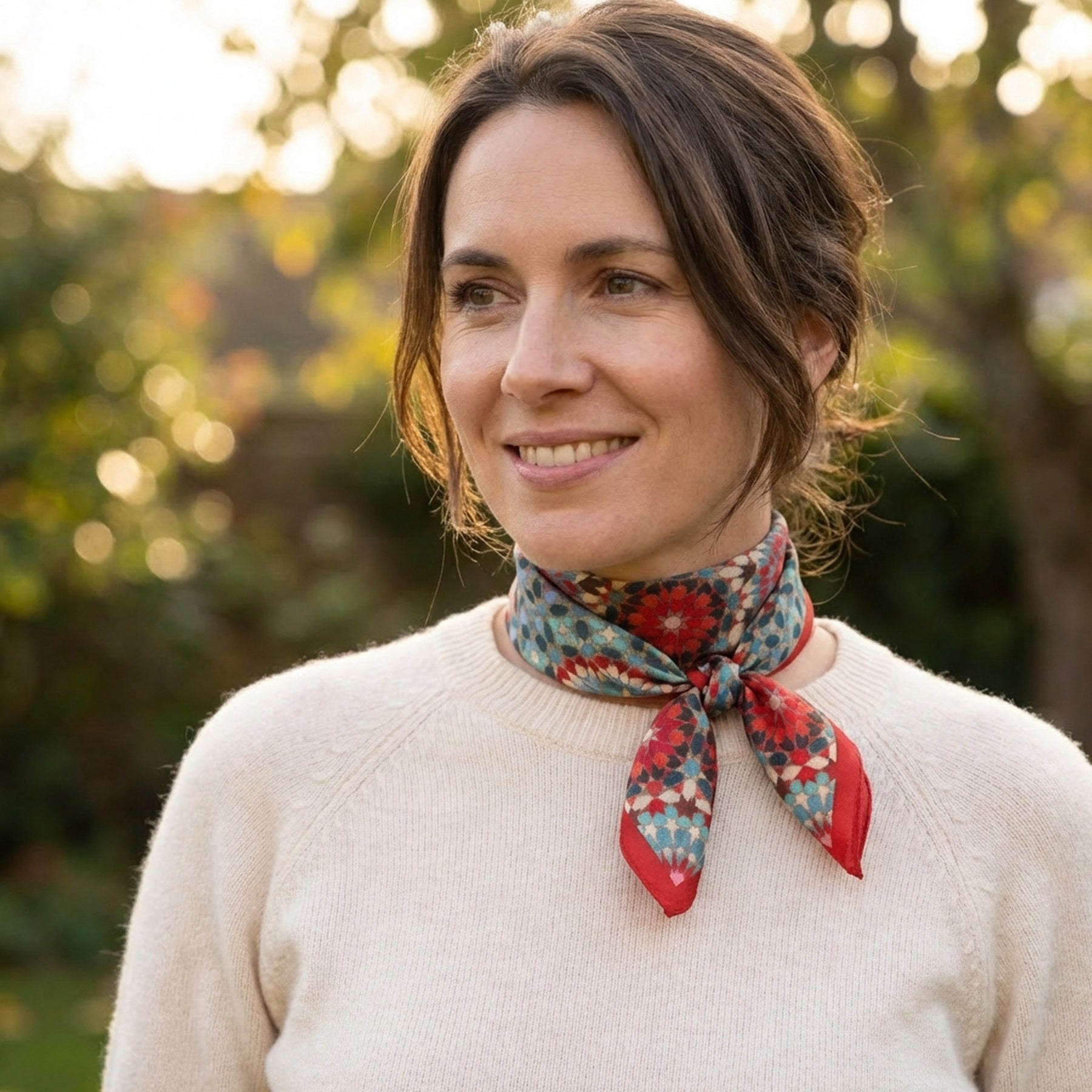 A woman with brown hair tied back smiles outdoors in front of blurred greenery, wearing a cream sweater and the Colorful Silk Neckerchief around her neck.