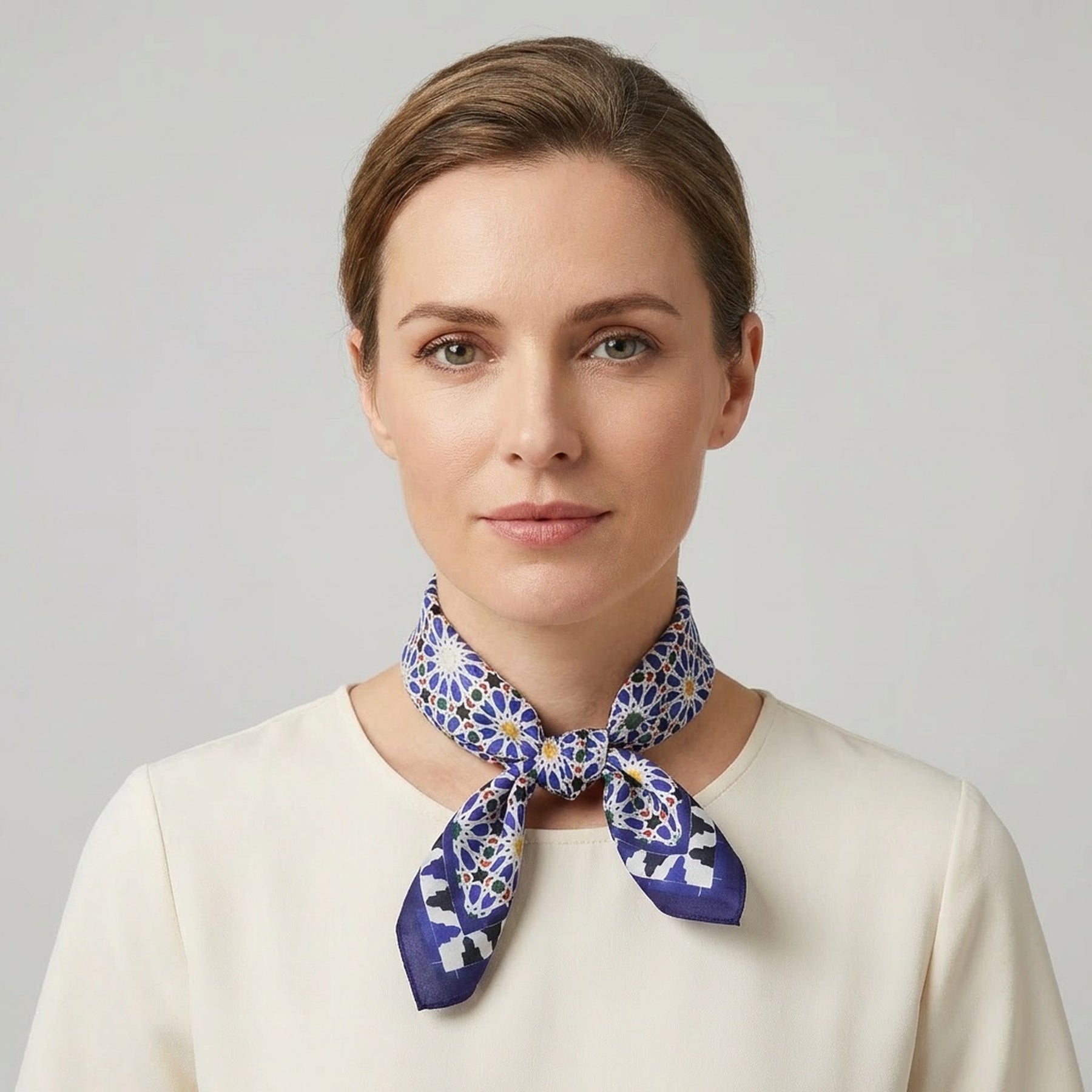 A woman with light brown hair in a neat bun wears a cream-colored top and the Blue Square Scarf Zellige v2 tied around her neck, standing against a plain light background with a neutral expression.