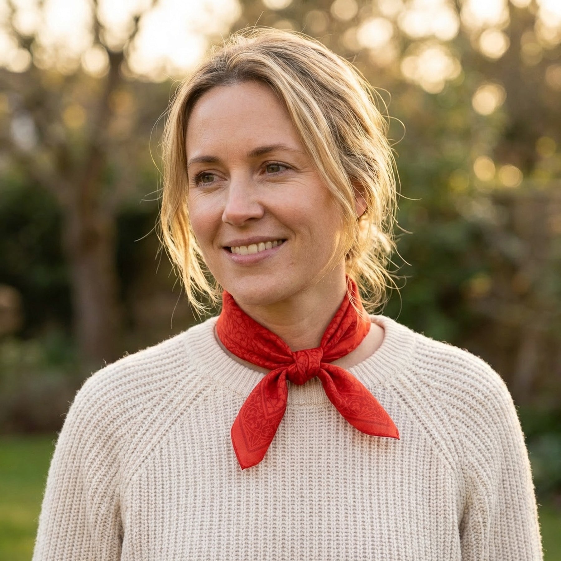 A woman with blonde hair in a loose bun, wearing a cream sweater and the Red Square Scarf Dalila v2 - 100% Silk tied around her neck, smiles while standing outdoors with greenery in the background.