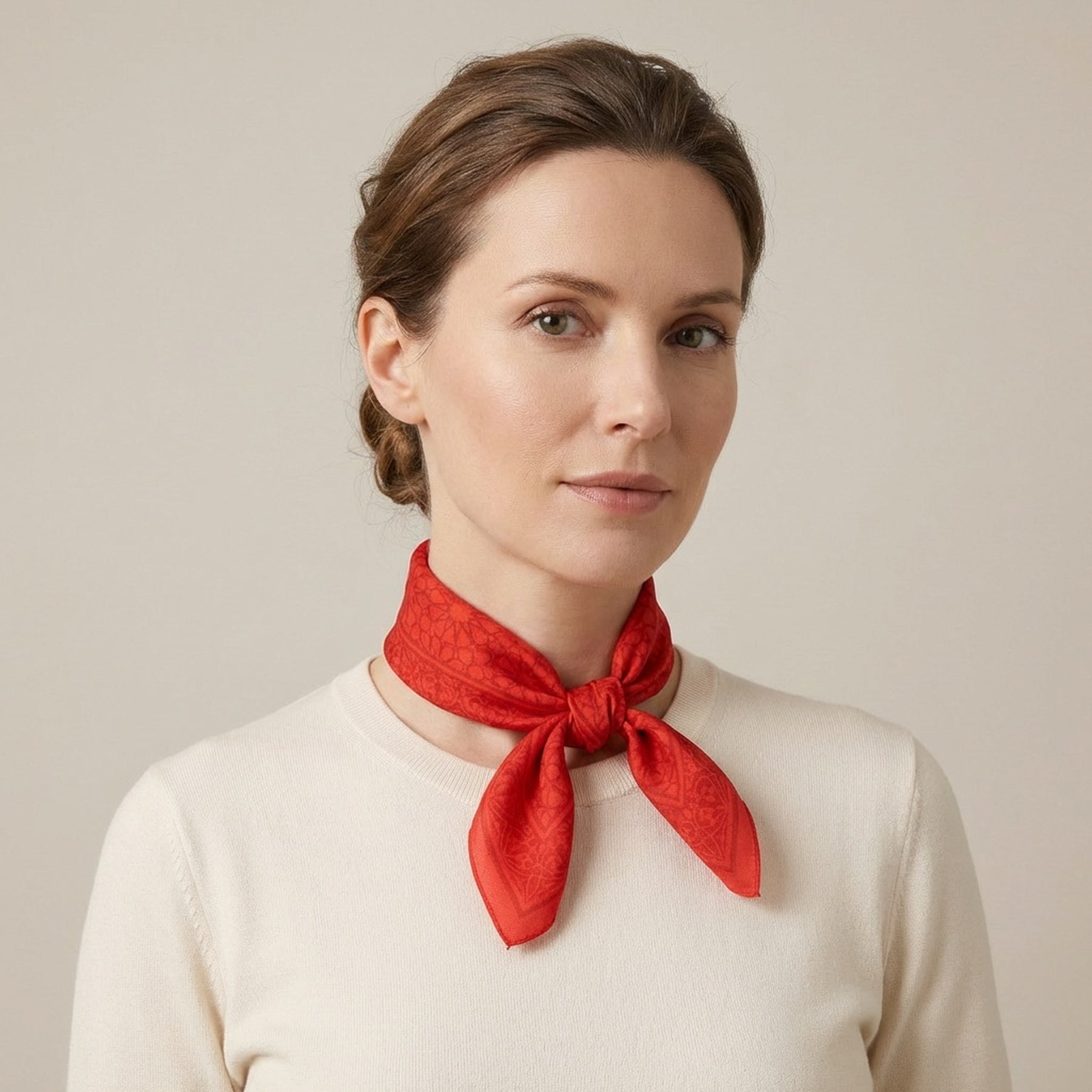 A woman with light brown hair in a low bun wears a cream sweater and the Red Square Scarf Dalila v2—100% Silk—neatly tied around her neck, standing against a plain beige background.