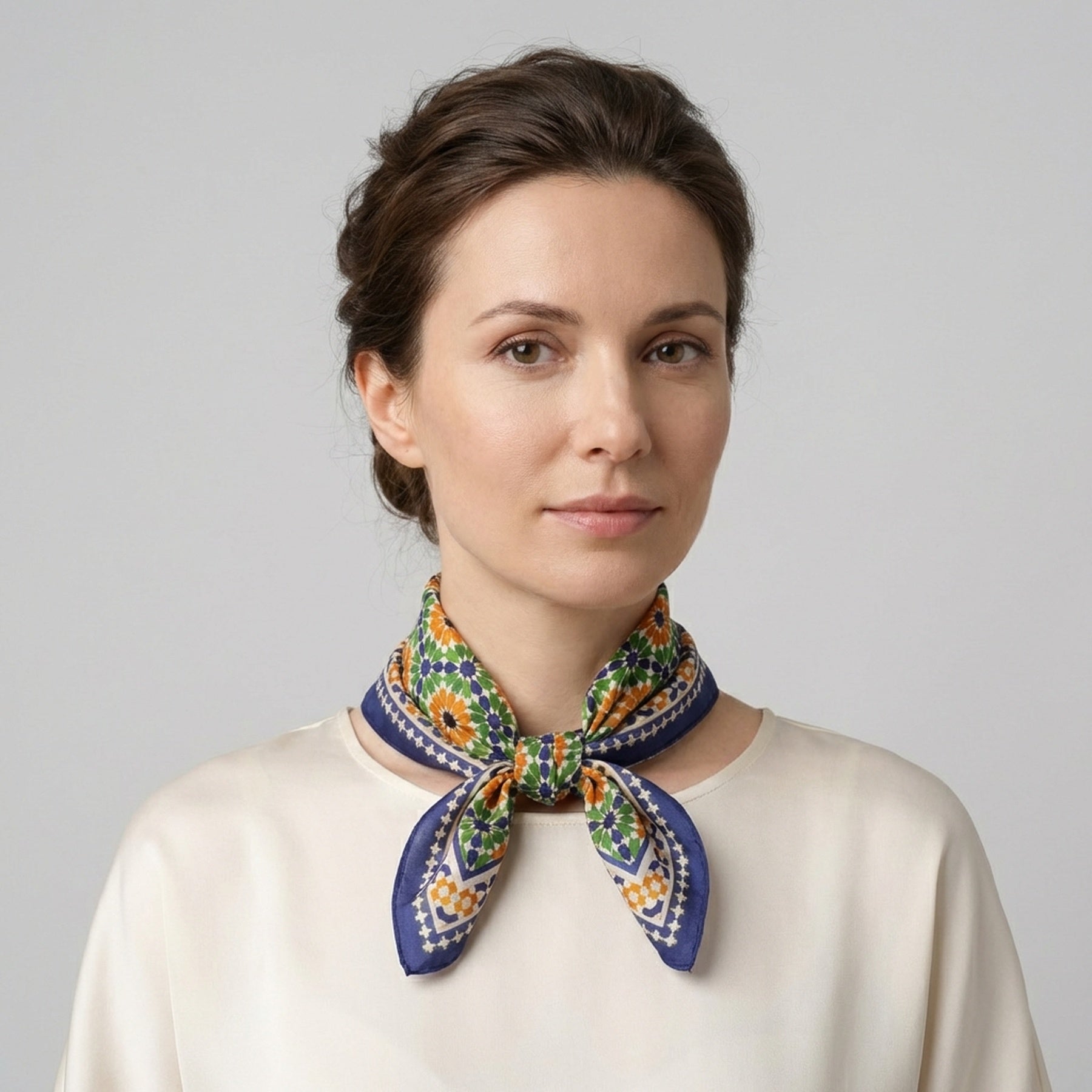 A woman with dark hair in an updo wears a cream blouse and the Islamic Art Print Square Silk Scarf Nur tied elegantly around her neck, standing against a plain light gray background.